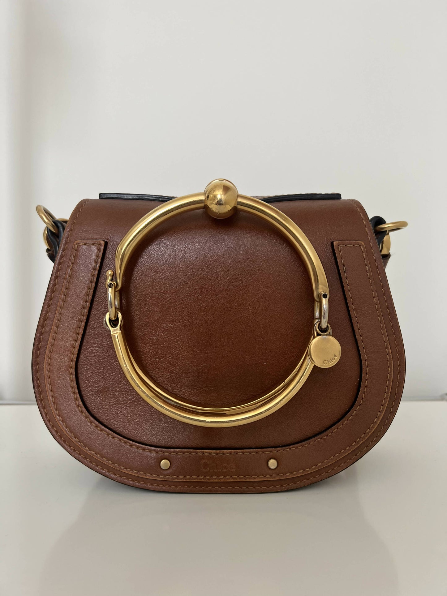 The EXCHANGE Chloe Nile Bag is a small brown leather designer handbag featuring a curved flap, decorative stitching, and a distinctive round gold handle and hardware, shown against a plain white background.