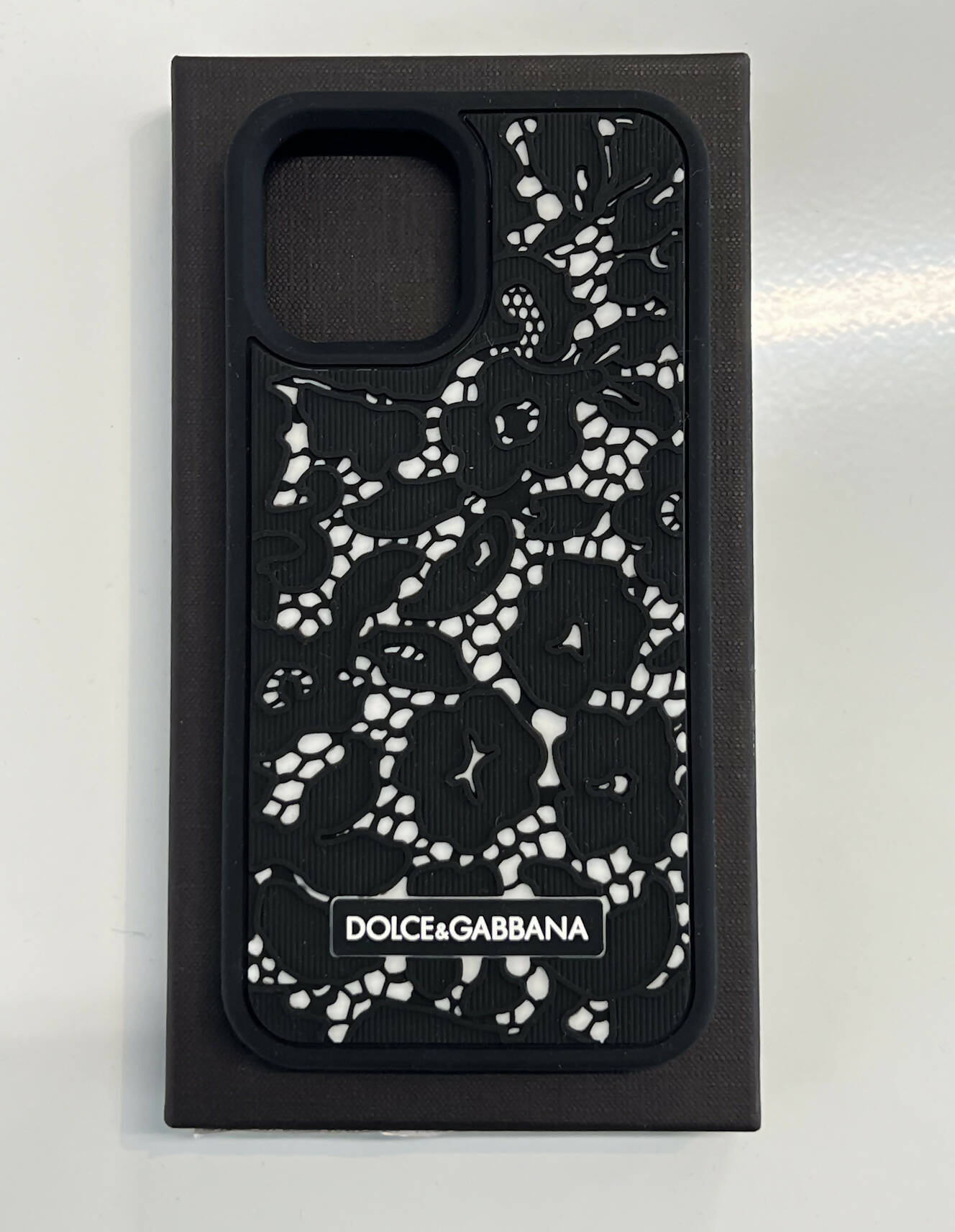 A Dolce & Gabbana - iPhone 13 Phone Case from EXCHANGE is shown in new condition with its box and dustbag, featuring a black design with white floral lace pattern atop a black rectangular box.