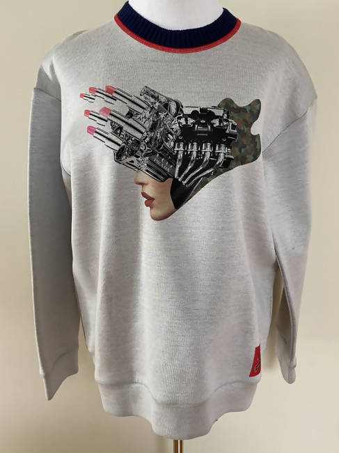On a mannequin, the PRADA 2X4 Collaboration Jumper by EXCHANGE in gray features a graphic of a womans face mixed with mechanical fingers. The navy neckline has a red stripe, and theres a small red tag at the lower side.