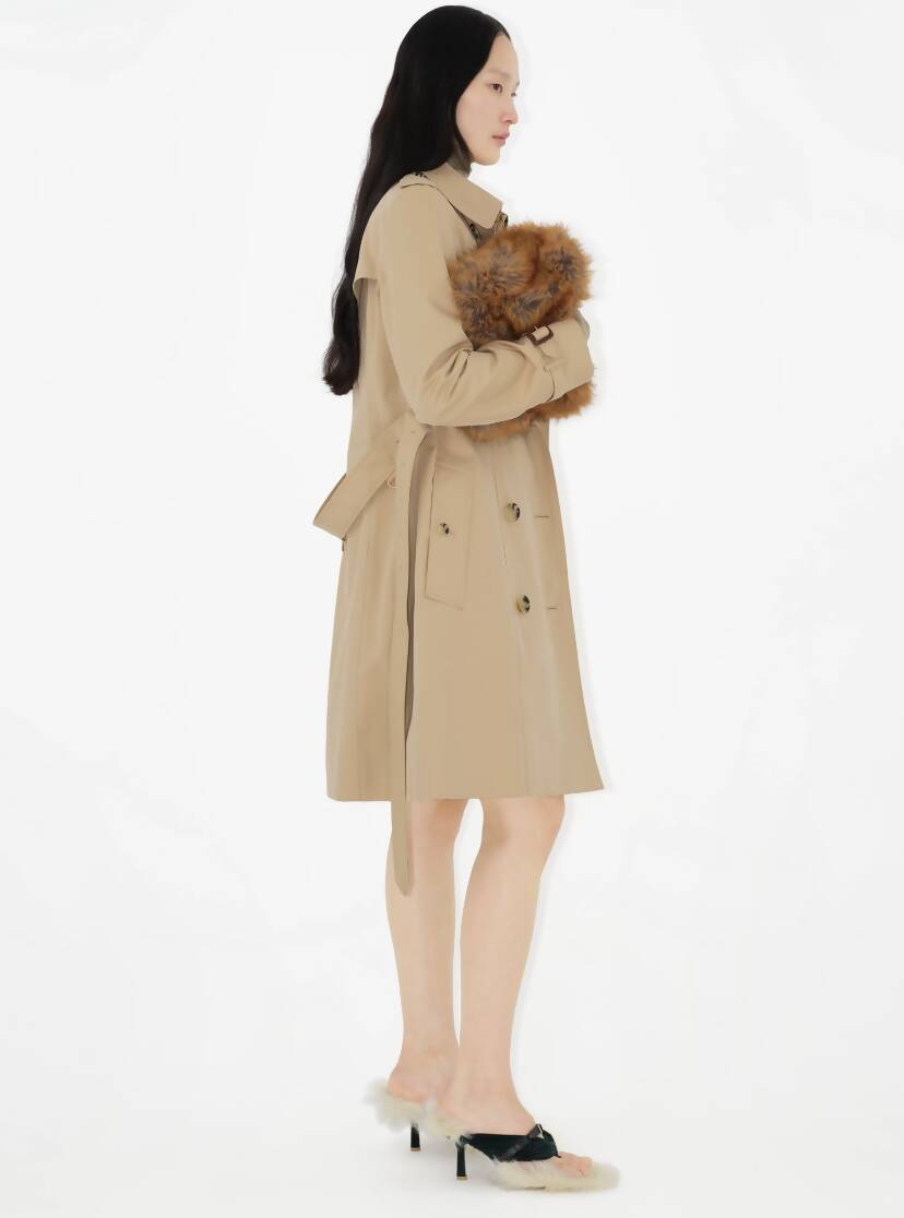 A woman with long dark hair in profile wears the EXCHANGE Burberry Mid-length Chelsea Heritage Trench Coat (UK Size 6), holding a small fluffy brown dog against a plain white background.