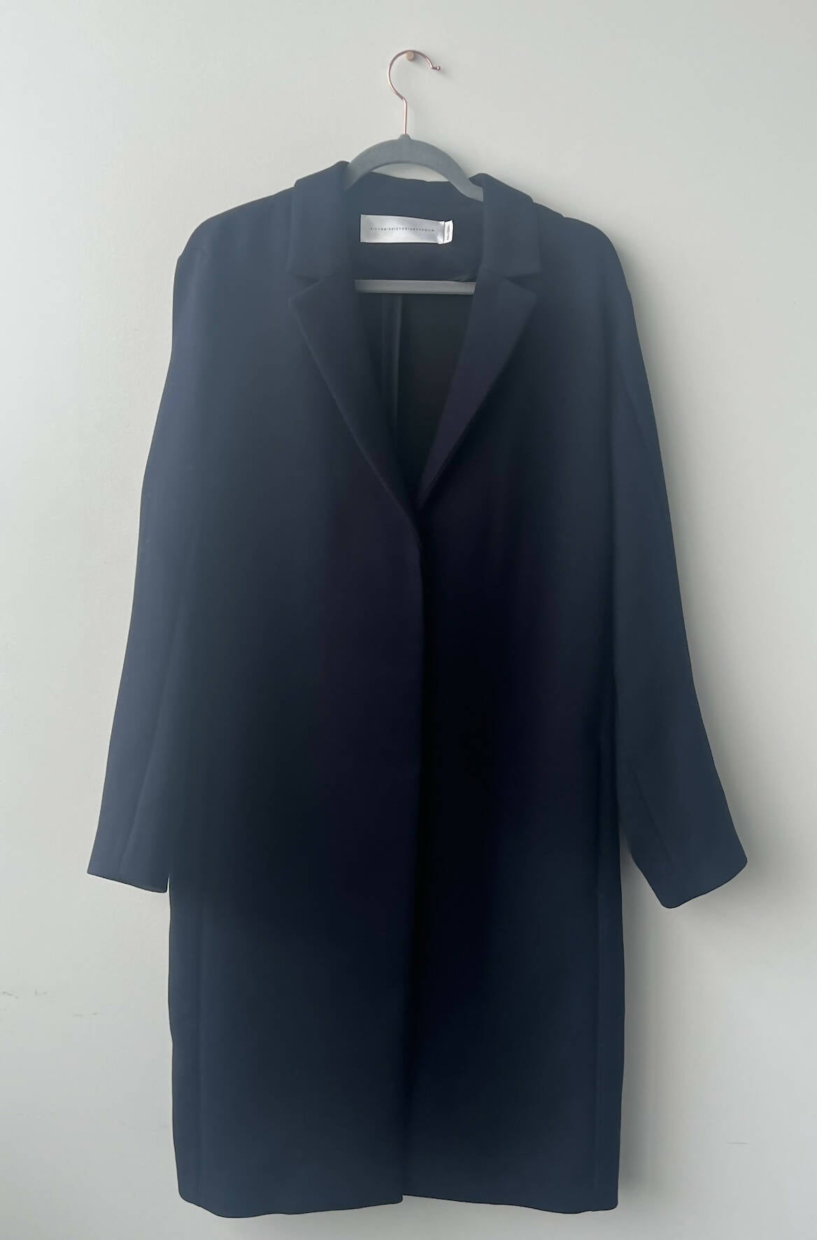 A navy blue Victoria Beckham coat with collar and lapels from EXCHANGE hangs on a black hanger against a light wall—an ideal find for any exchange-store shopper.