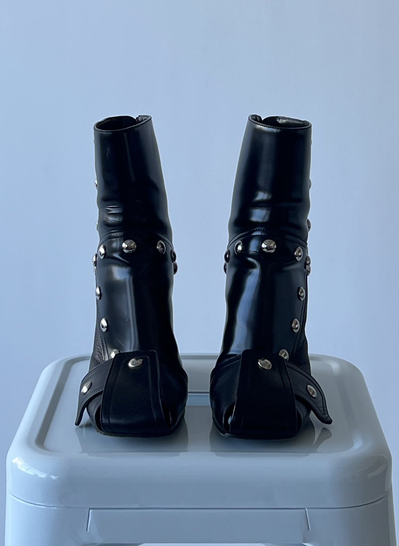 A pair of black, shiny, high-heeled boots with silver studs and square toes from the EXCHANGE LV Fall Winter 2021-2022 - Full Set rests on a white surface against a plain light blue background.