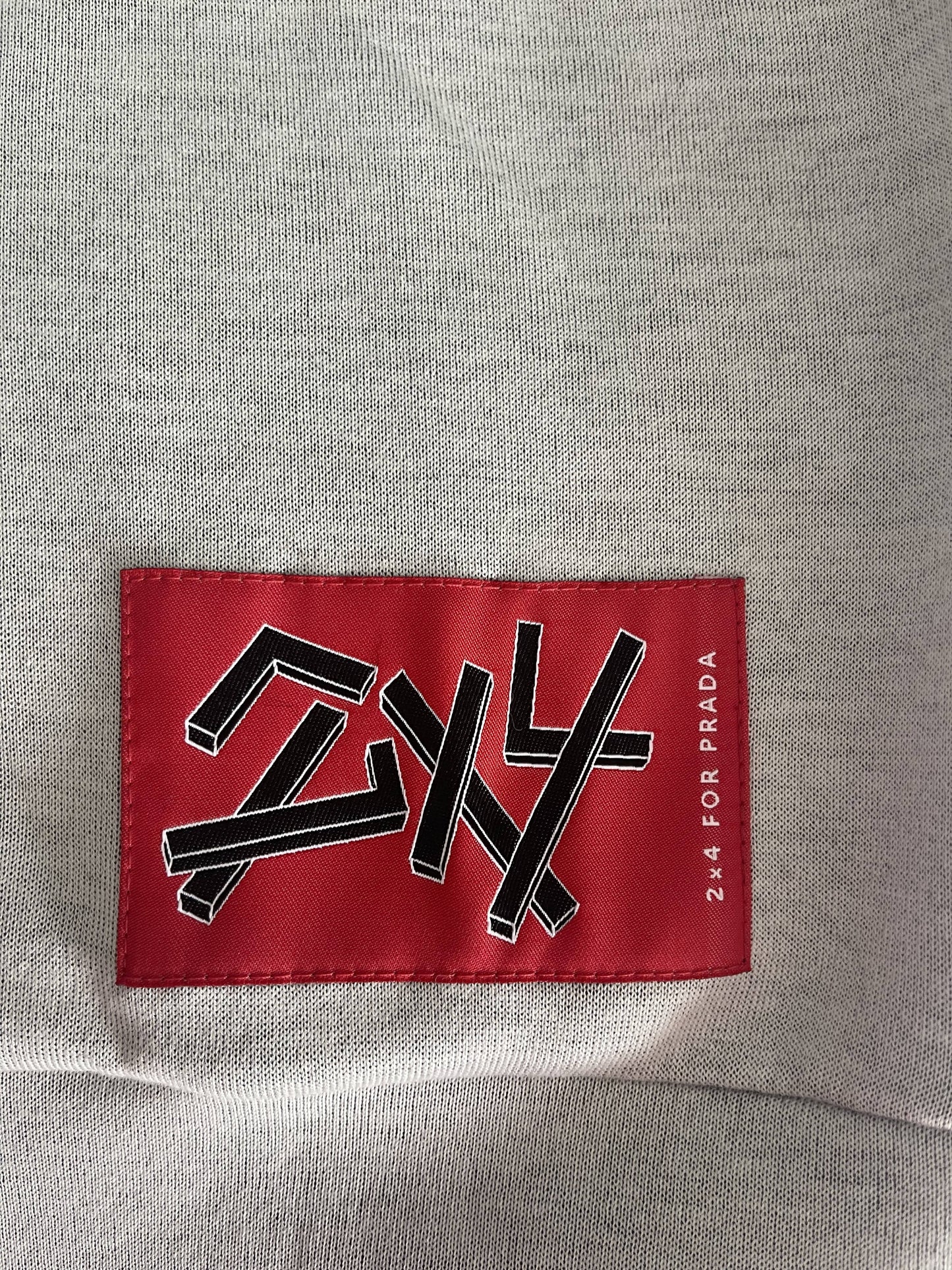Close-up of a gray fabric with a red patch sewn onto it, featuring a lipstick face logo. The black and white stylized text resembles Japanese characters and reads 24 4 FOR PRADA on the side, part of the EXCHANGEs PRADA 2X4 Collaboration Jumper, Grey.