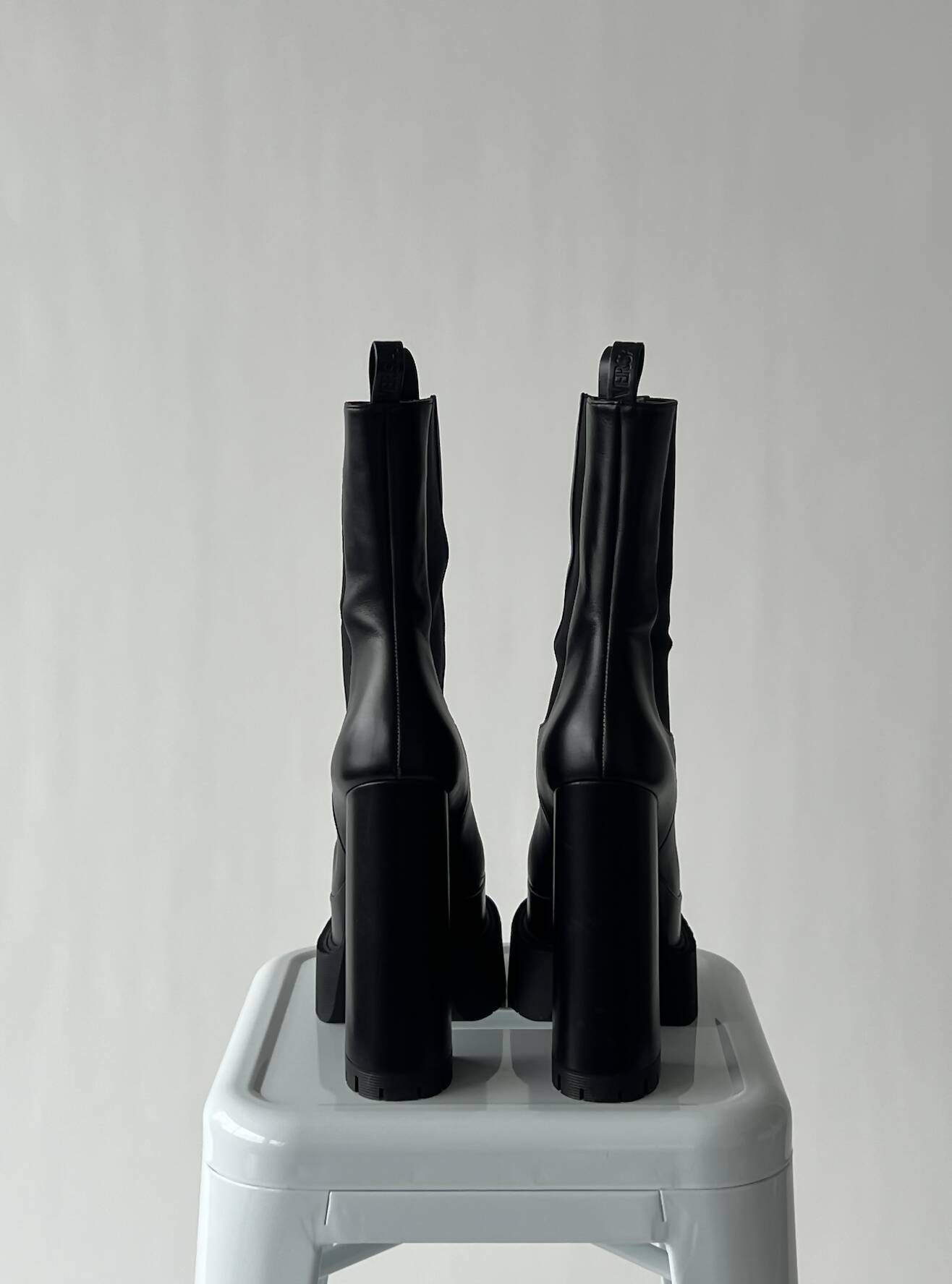 A pair of black VERSACE Aevitas Platform Boots - FULL SET by EXCHANGE, size 39, is shown from the back, side by side on a light gray plastic stool against a plain light background.