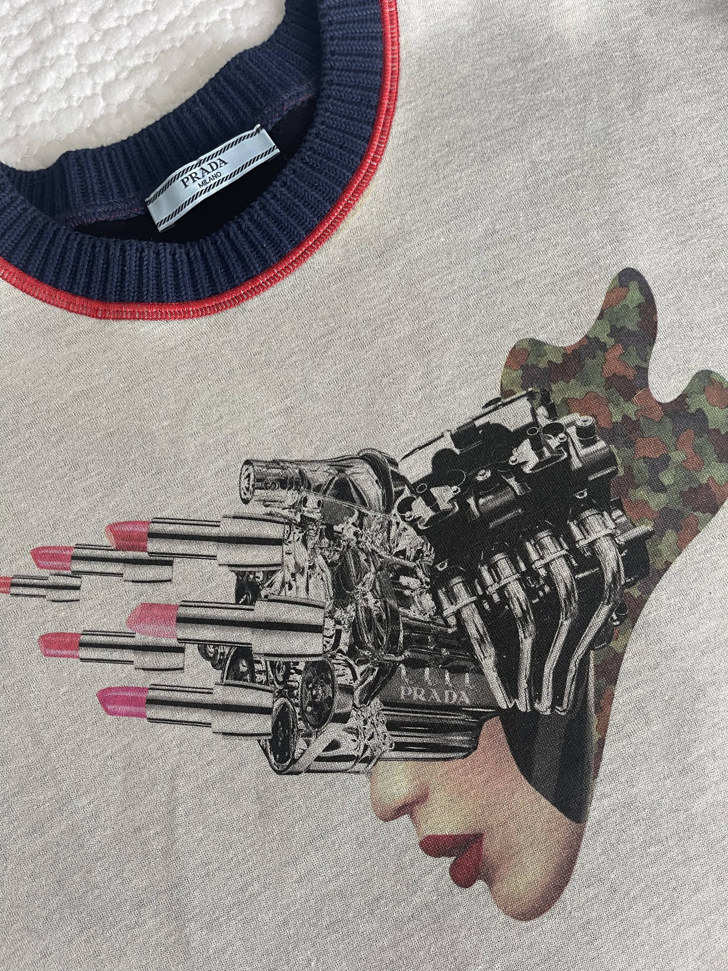 A close-up of the PRADA 2X4 Collaboration Jumper by EXCHANGE in gray showcases abstract designs with lipstick tubes, metallic machinery, and a camo-patterned face. The navy neckline with red trim provides striking contrast.
