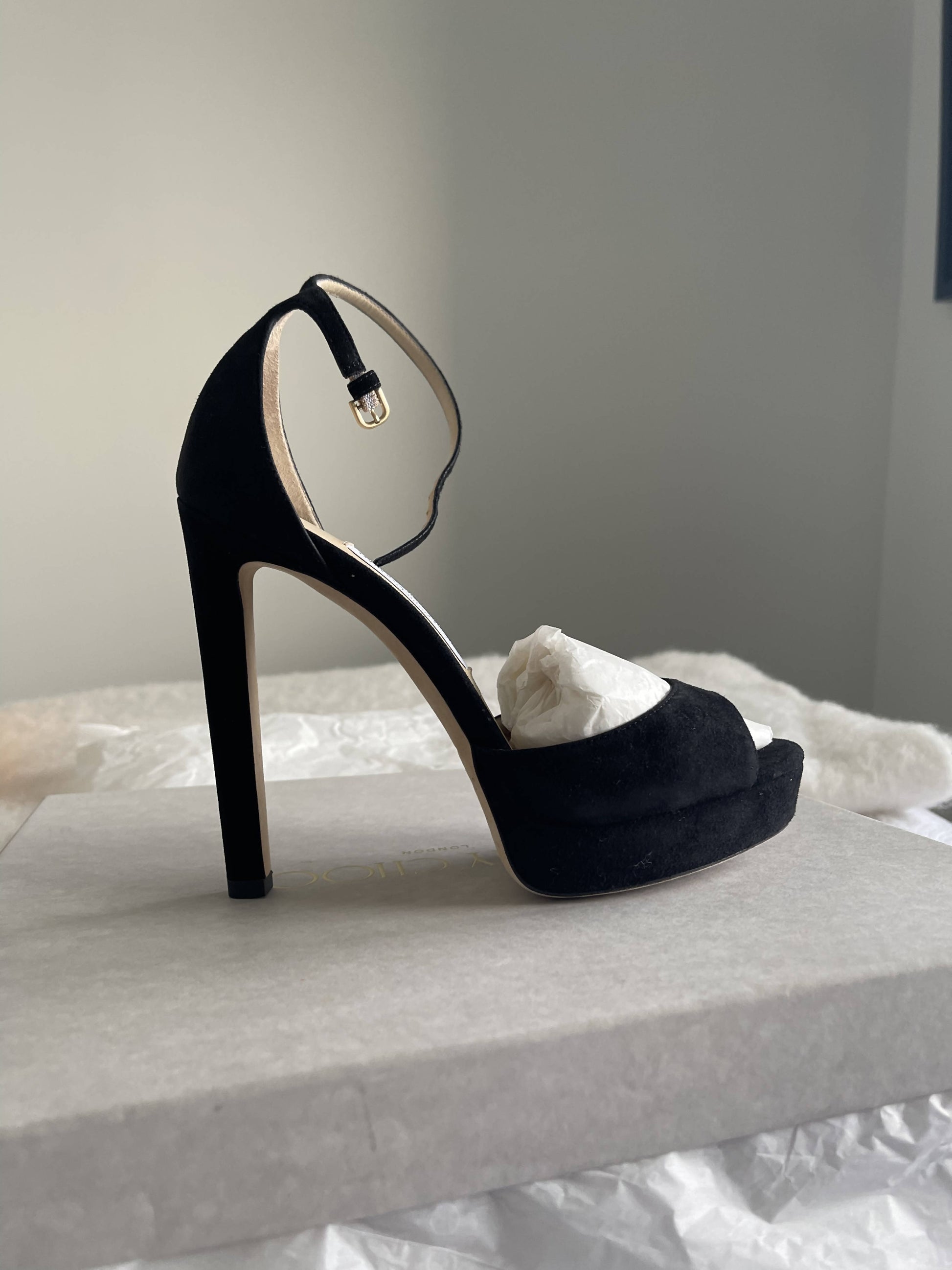 A black EXCHANGE Jimmy Choo Pattie Heel, size 37.5, open-toe platform with ankle strap and buckle, sits on a gray box filled with tissue paper. The softly lit neutral background highlights its excellent condition.
