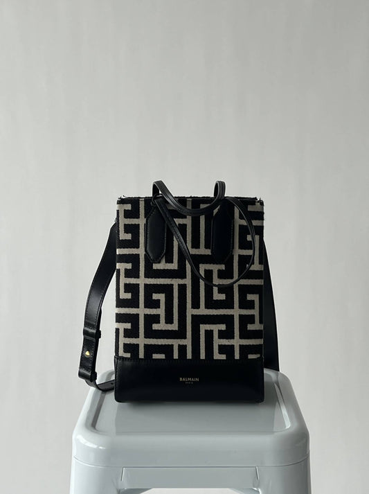 A BALMAIN Maxi Monogram Jacquard Shopping Tote by EXCHANGE, featuring a black and beige geometric pattern with black leather accents and a gold-tone logo charm, rests on a white stool against a light gray background.