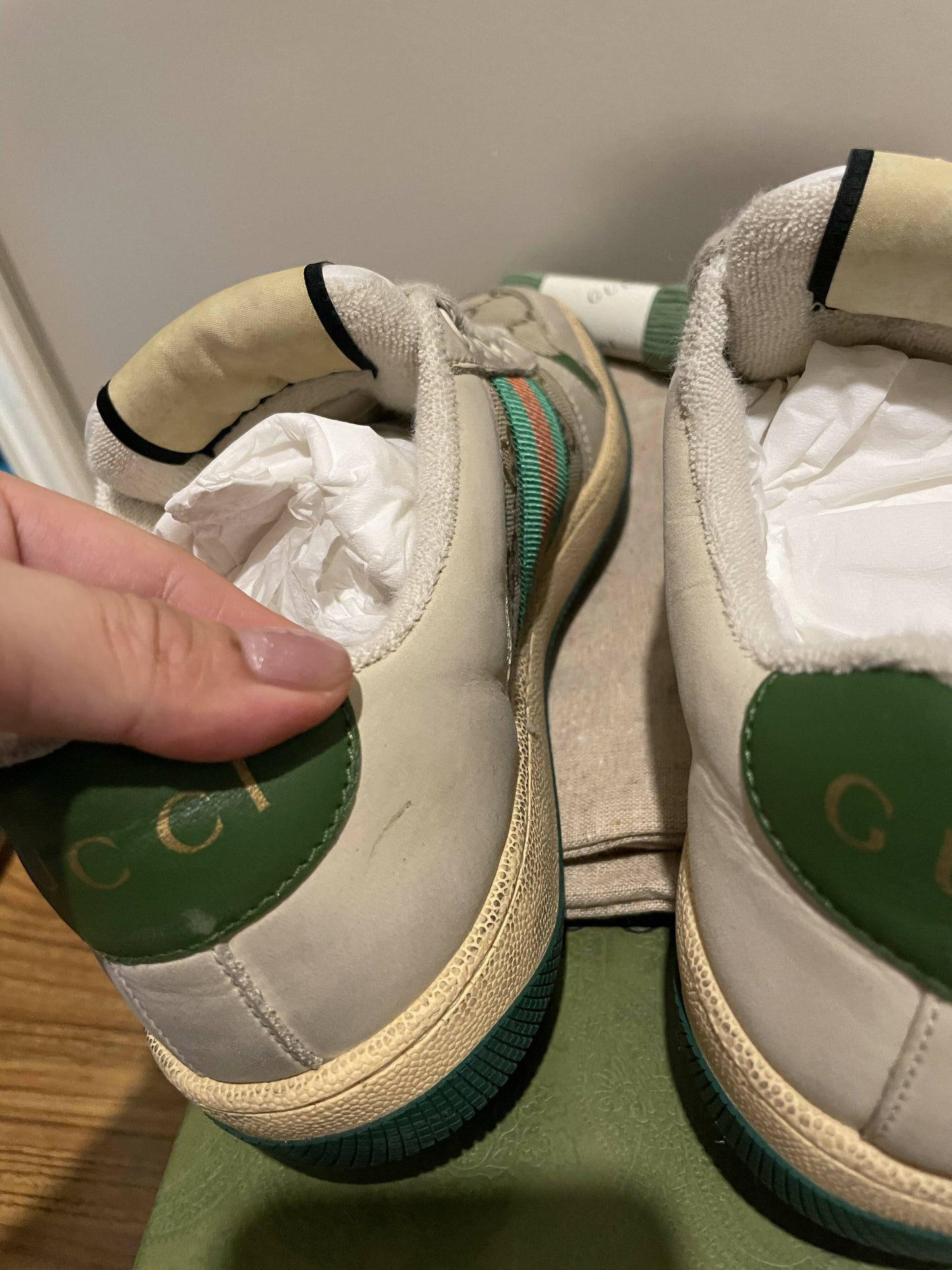 Close-up of two white EXCHANGE Gucci Women's Screener leather sneakers with green and red stripes, gold GUCCI lettering on green heel tabs, and paper inserts. A hand holds one shoe on a green surface, emphasizing their vintage sportswear style.
