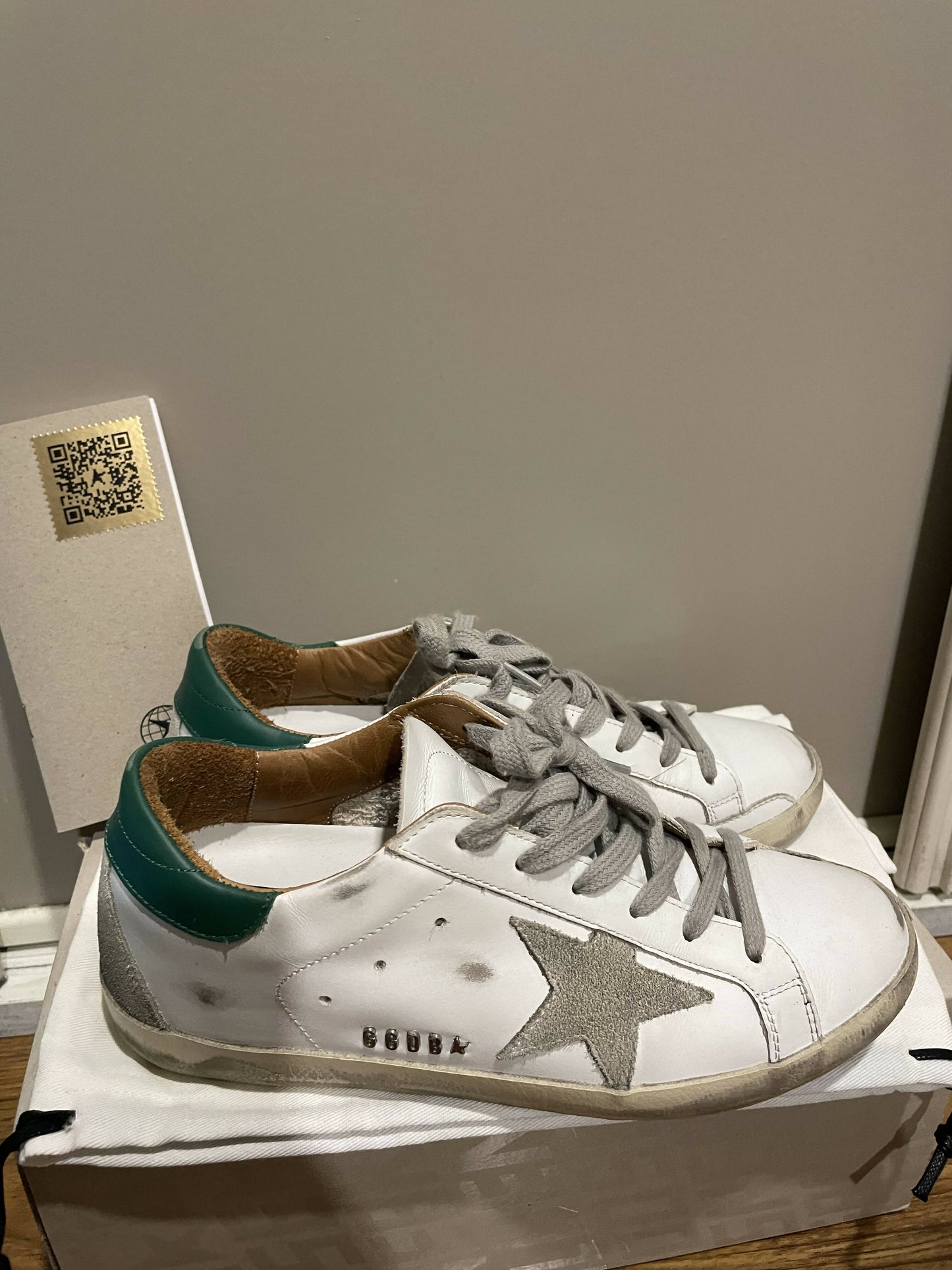The Golden Goose Superstar sneakers by EXCHANGE feature gray laces, a gray star design, and green heel accents in Italian leather, displayed on a white shoebox beside a wall with a visible QR code.