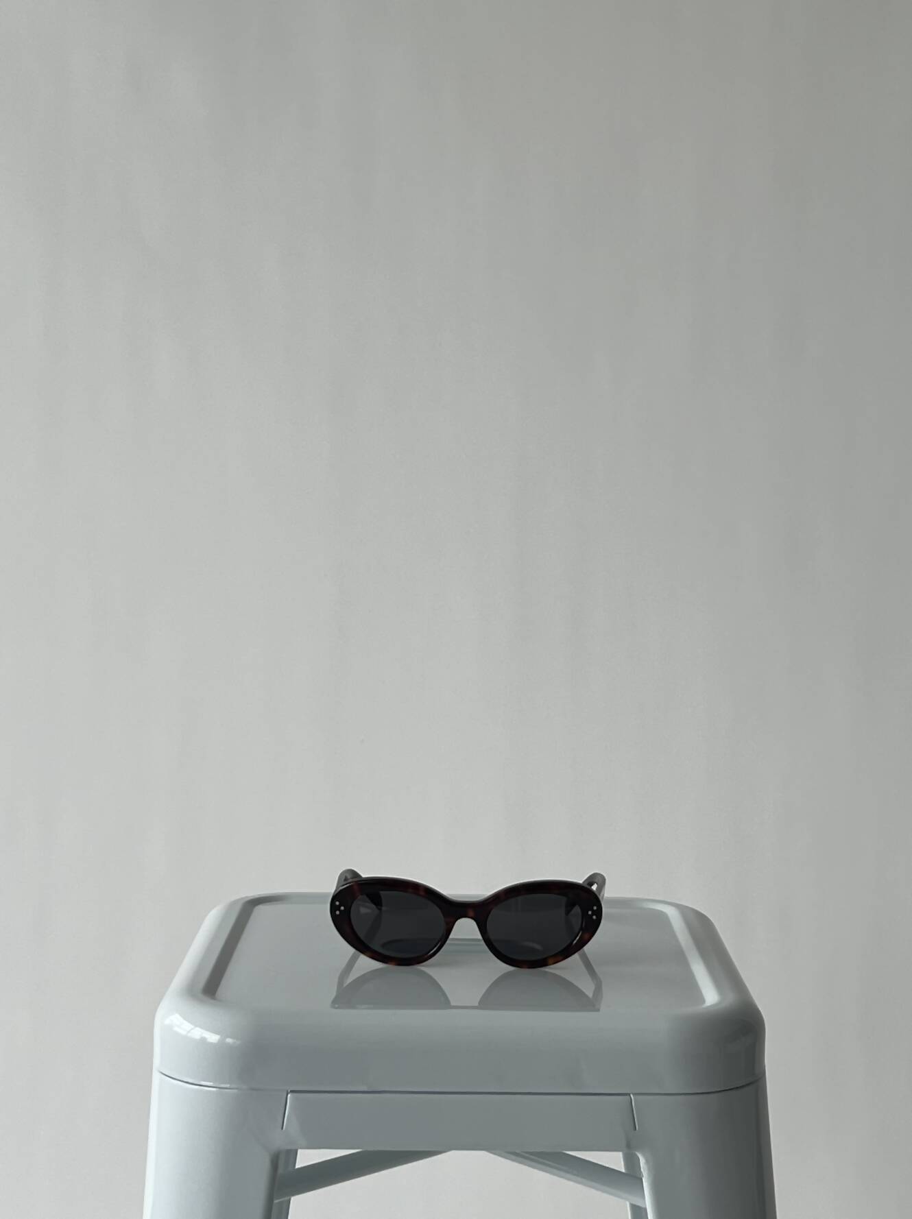 A pair of EXCHANGE Celine Glasses, worn once, rests on a light gray plastic stool against a plain, light-colored background.