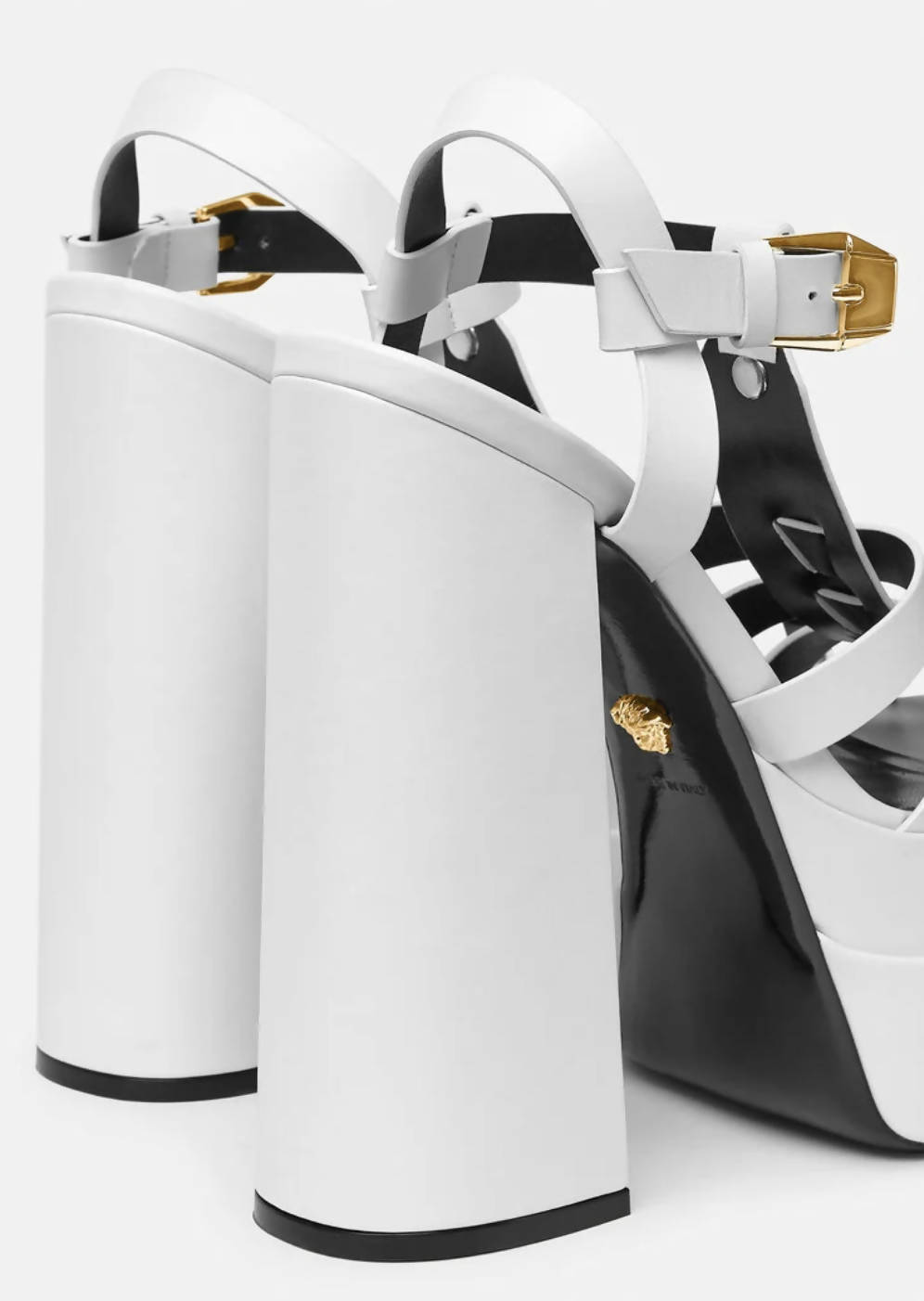 Close-up of LA MEDUSA PLATFORM SANDALS by EXCHANGE featuring leather slingback design, thick white block heels, black soles, white straps, gold-tone buckles, Medusa hardware, and a gold embellishment near the heel.