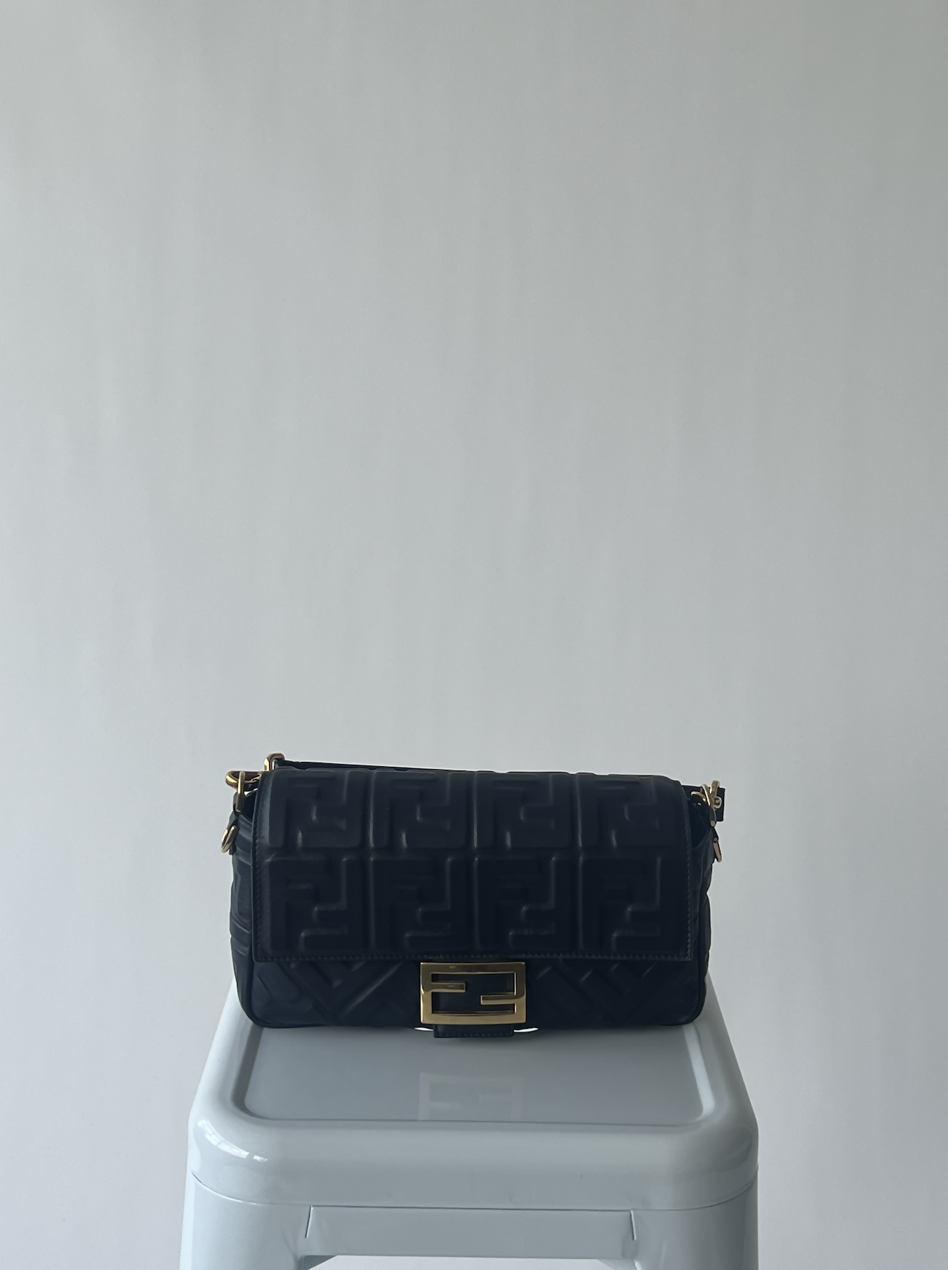 A black EXCHANGE Fendi Nappa Embossed FF 1974 Baguette handbag with a gold clasp and geometric pattern rests on a white stool against a plain light gray background.