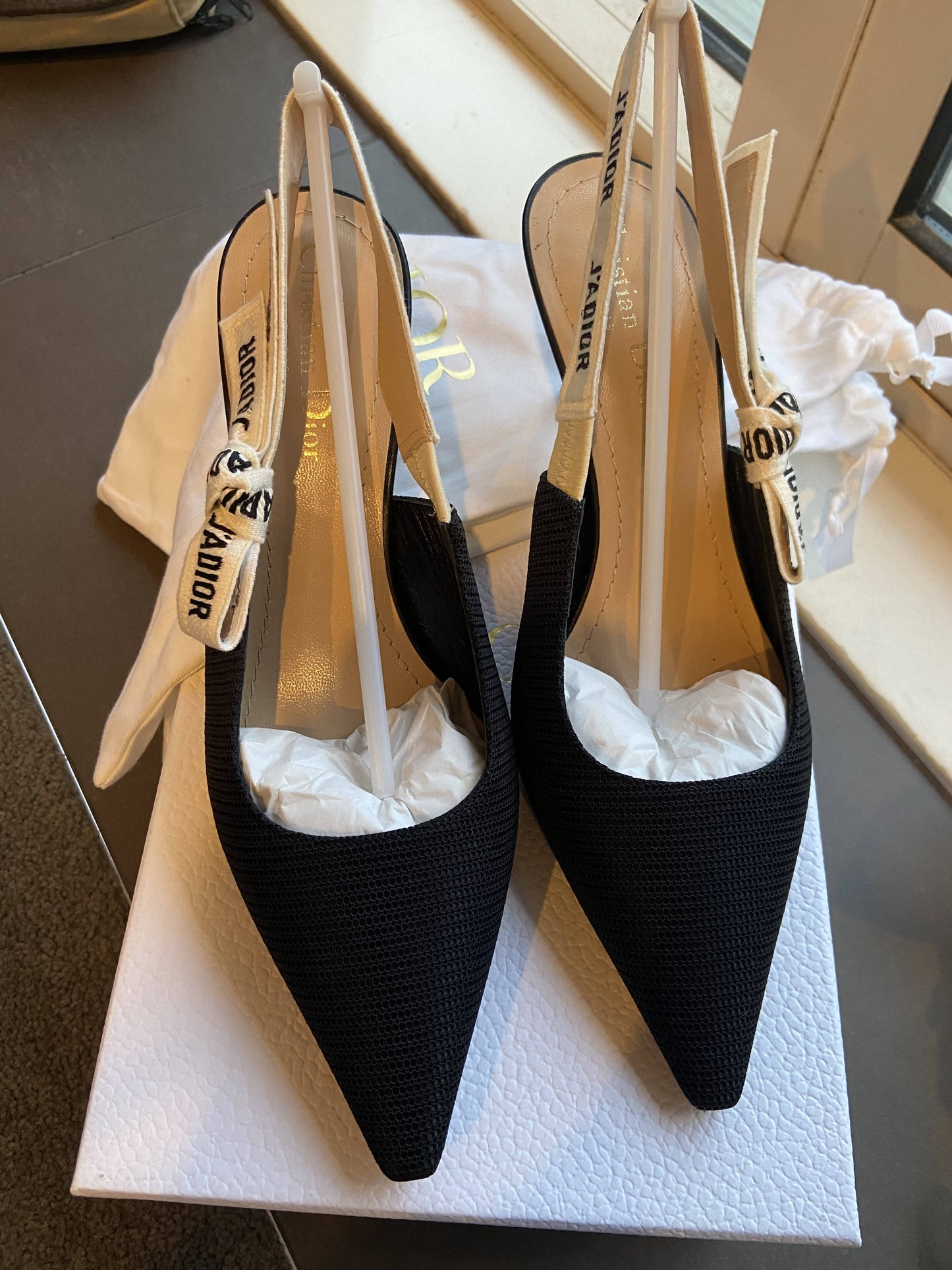 A pair of EXCHANGE Dior J’adior Slingback heels in size 37.5 with black pointed toes and white JADIOR ribbon straps, shown on a white box by a window.