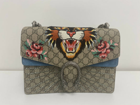 The EXCHANGE Dionysus Embroidered Tiger Medium Shoulder Bag features a roaring tiger face, pink hand-embroidered floral appliqué, GG beige patterned background, blue suede detail, and a silver snake-design clasp.