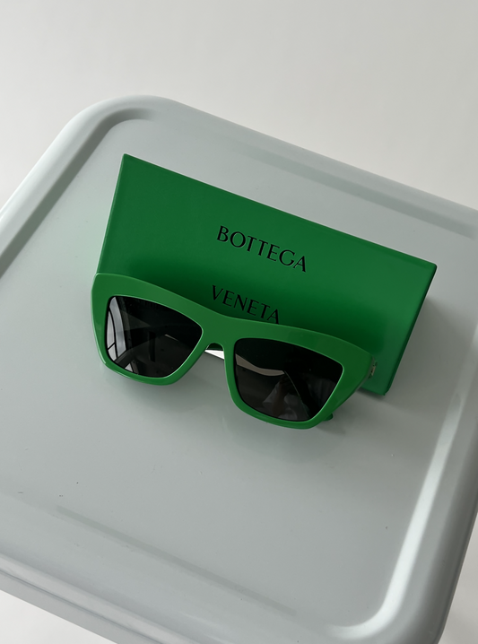 The Bottega Veneta New Entry Cat Eye Sunglasses by EXCHANGE, featuring dark lenses, are displayed on a matching green case atop a light gray surface.