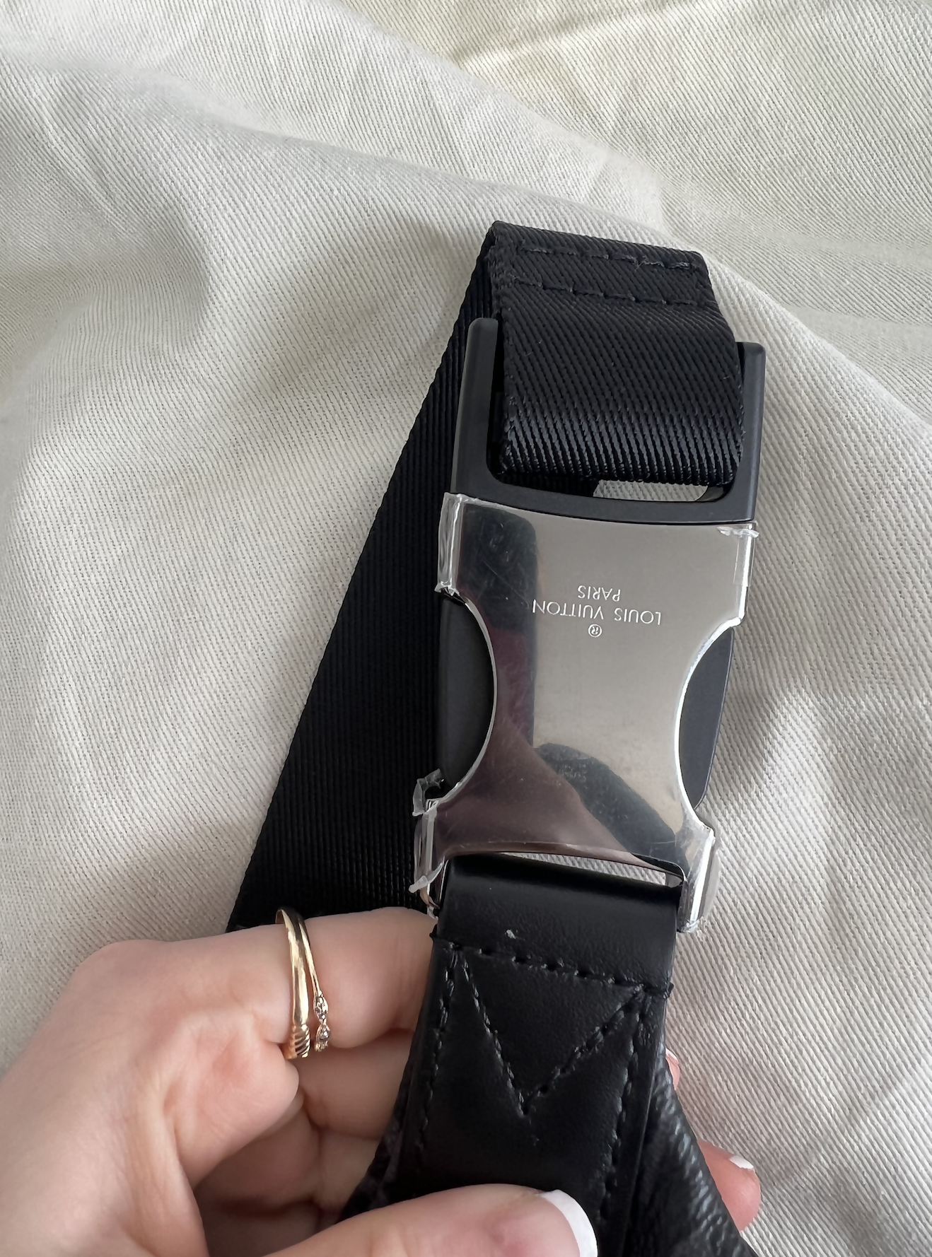 A hand with rings holds a pre-owned Louis Vuitton Discovery Bumbag Monogram Eclipse Black by EXCHANGE, set against a light fabric background.