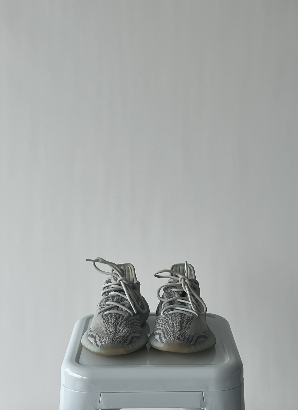 A pair of EXCHANGE Yeezy 350 - Blue Tint sneakers with gray and white patterned uppers and white laces sit side by side on a light gray stool against a plain light background.