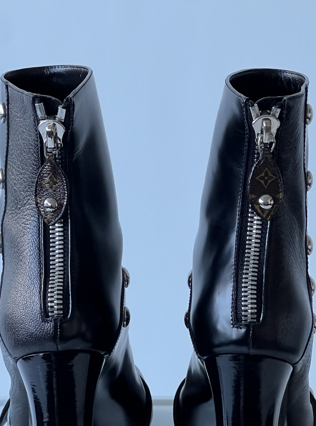Close-up of two black leather EXCHANGE ankle boots from the LV Fall Winter 2021-2022 - Full Set, featuring shiny silver zippers with round pull tabs embossed with a pattern, photographed against a pale blue background. Limited edition.