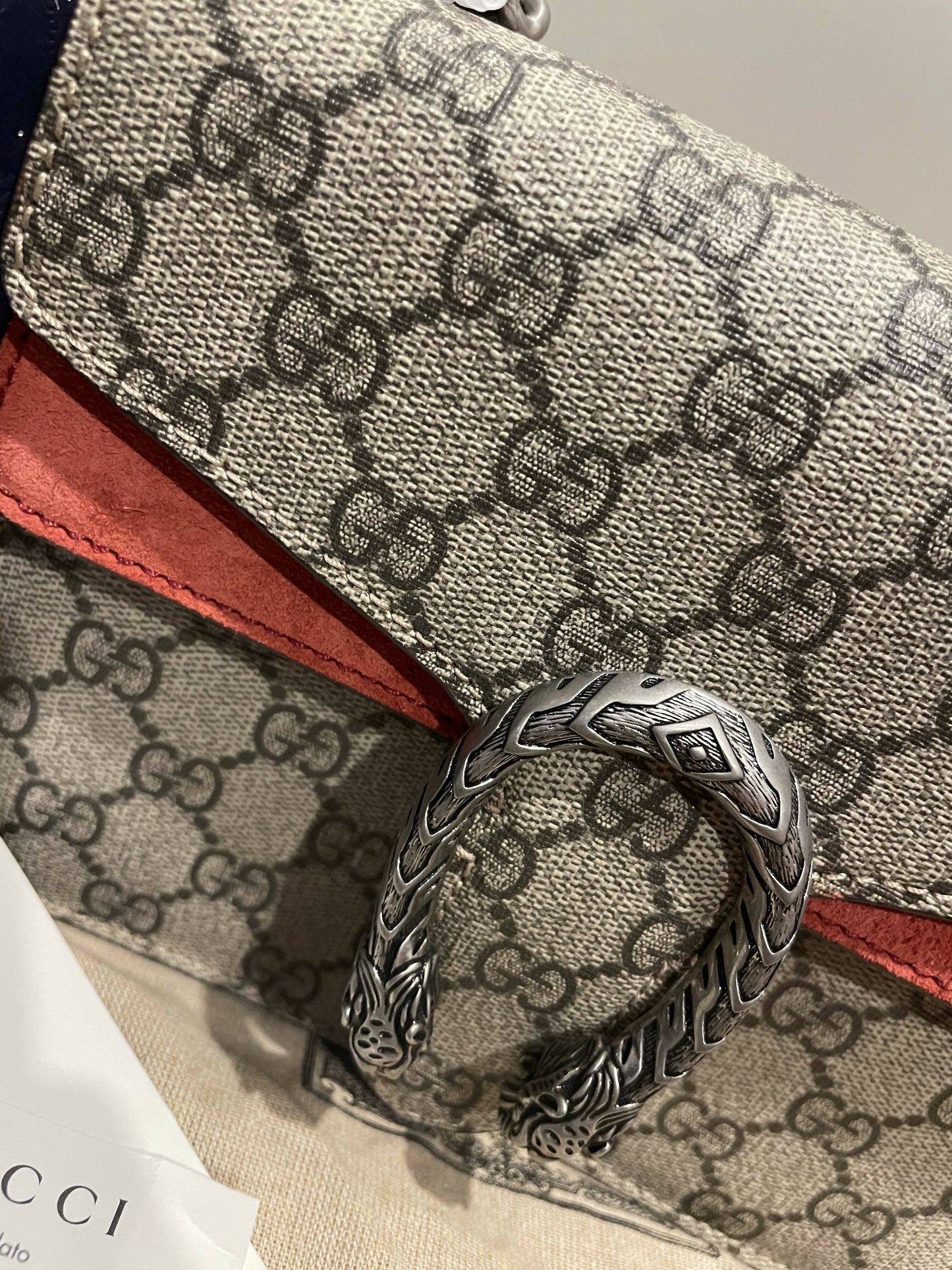 Close-up of an EXCHANGE Gucci Dionysus GG Supreme mini bag with a textured silver tiger head spur buckle and red suede details. Part of a Gucci tag is visible in the bottom left corner.