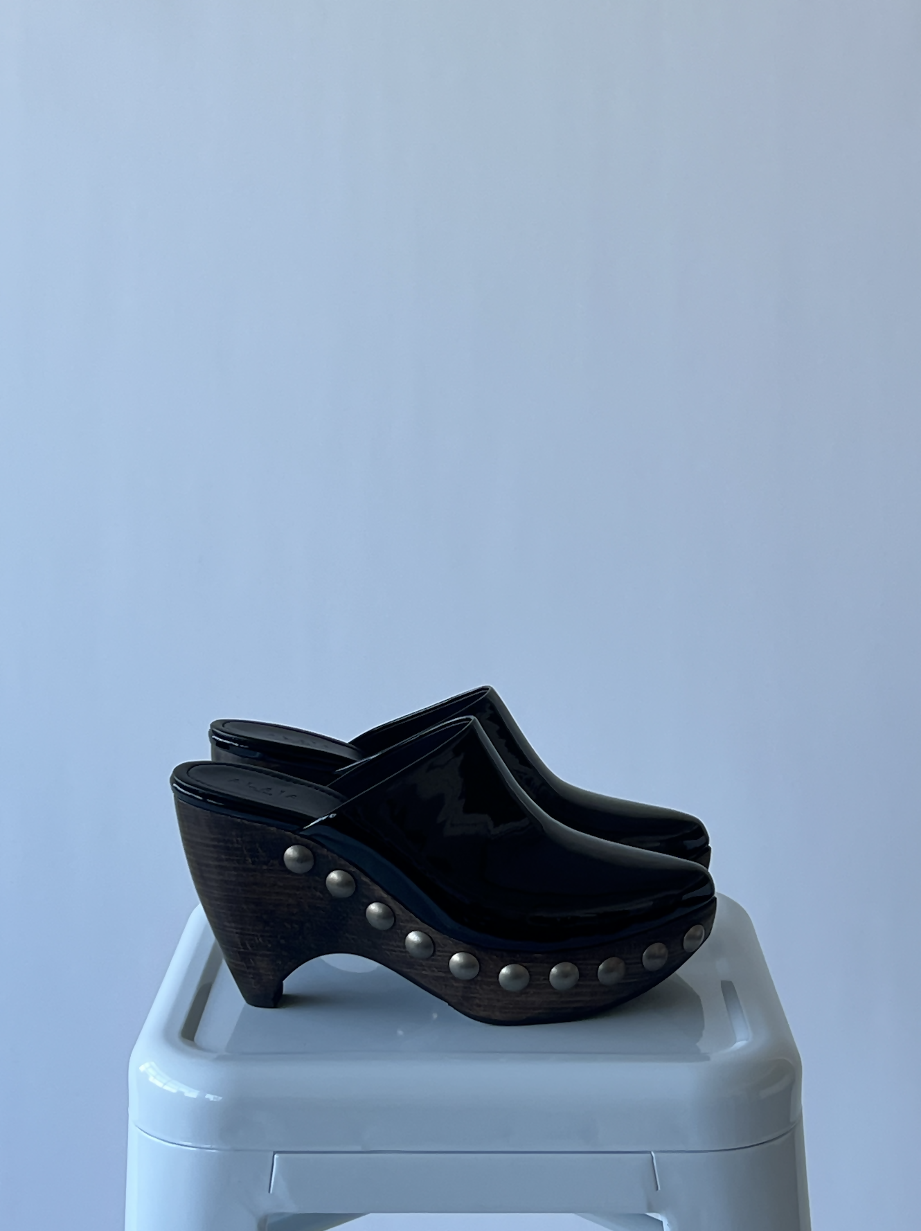The Alaïa Le Sabot leather and wood pumps by EXCHANGE, featuring a glossy black platform design with metal studs, are displayed atop a white stool against a plain light gray background.