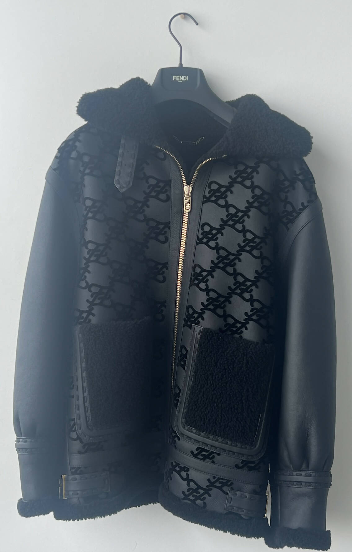 A Fendi Jacket Black shearling jacket by EXCHANGE features a monogram pattern, fuzzy collar, large front pockets, and black lamb leather sleeves—an effortlessly chic oversized aviator displayed on a branded hanger against a light gray wall.