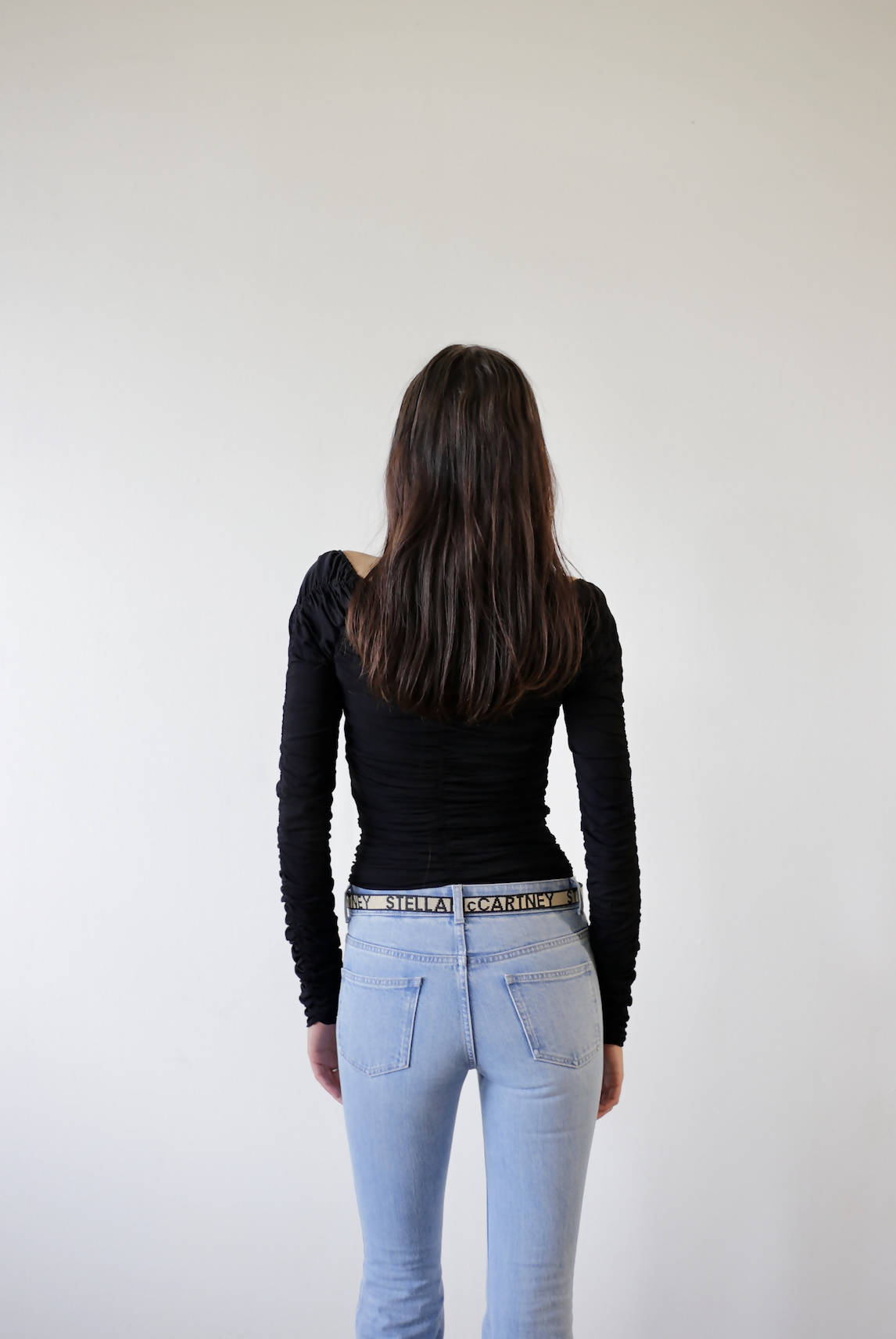 A person with long brown hair, wearing a fitted black Khaite Body Suit by EXCHANGE (Size 6) and light blue jeans, stands facing a blank white wall with their back to the camera.