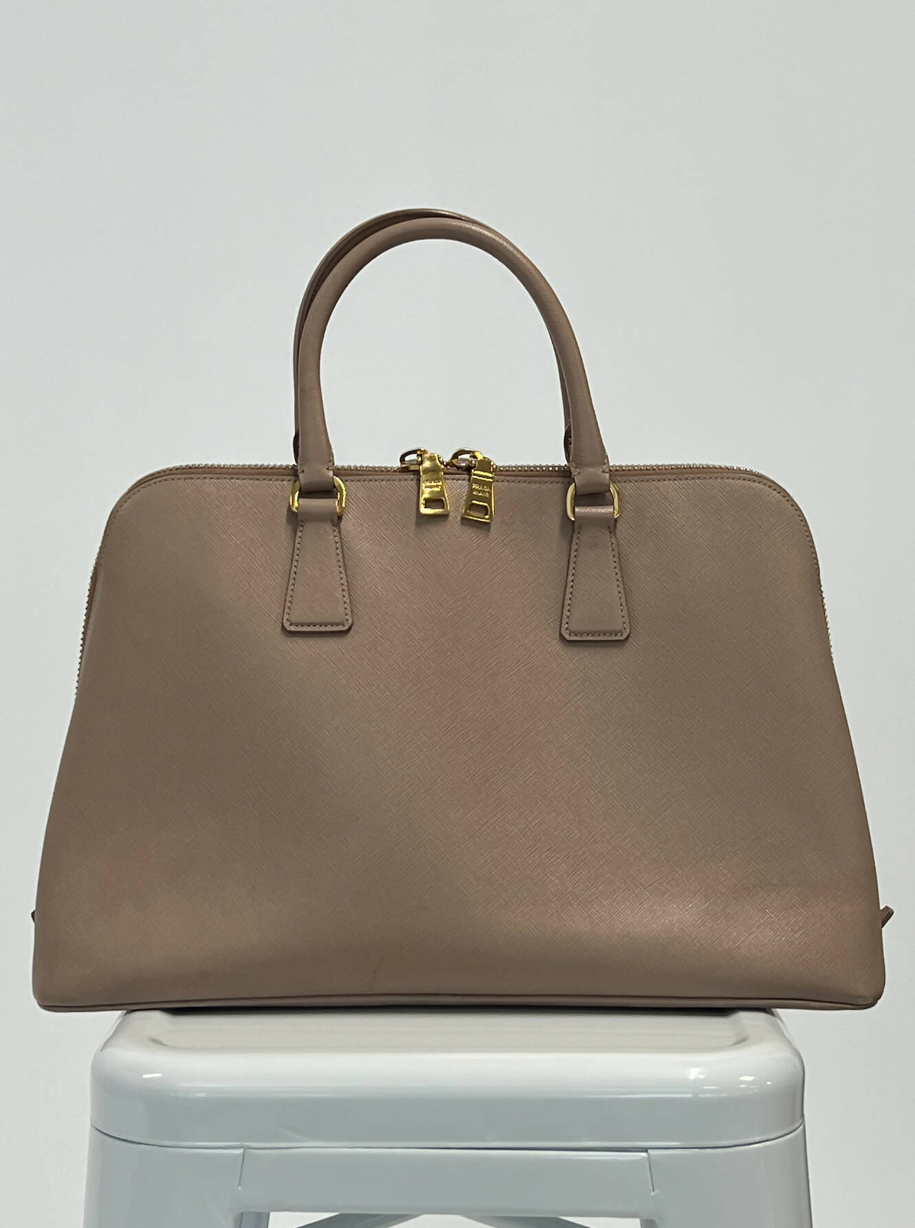 The EXCHANGE Prada Handbag, featuring taupe leather, gold hardware, and double handles, is displayed on a white stool with its dust bag against a plain white background.