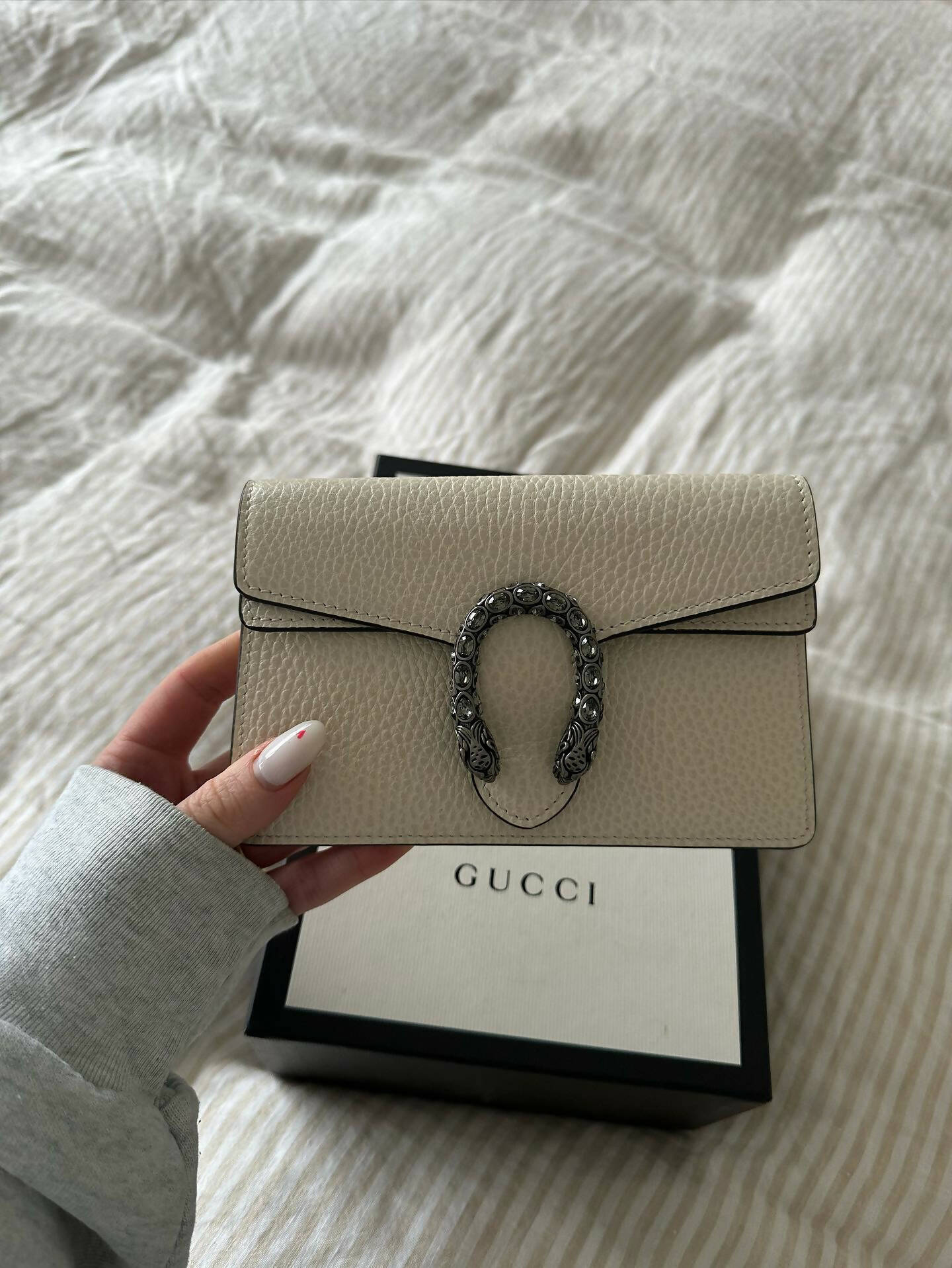 A hand holds an EXCHANGE Gucci Dionysus Super Mini Crossbody Bag – FULL SET, crafted in Italy from beige textured calf leather with a silver horseshoe clasp, above a Gucci box on a striped bedspread.