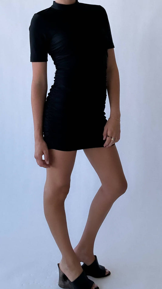 A person models the EXCHANGE Alexander Wang Rushed Dress, a fitted black short-sleeve medium dress, with black open-toe heeled sandals against a plain light background. Their left knee is bent and their hand rests at their side.
