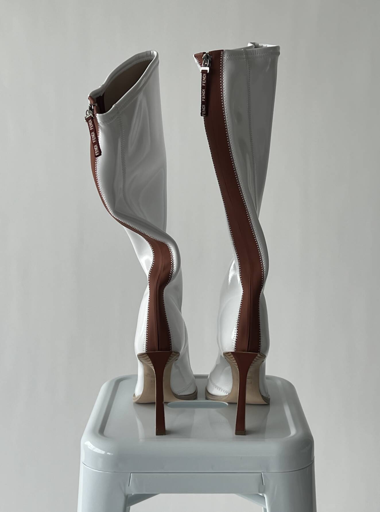 A pair of EXCHANGE FENDI Glossy Neoprene FFrame Boots, size 40, featuring bold wavy brown back zippers, stands on a white stool against a plain light gray background.