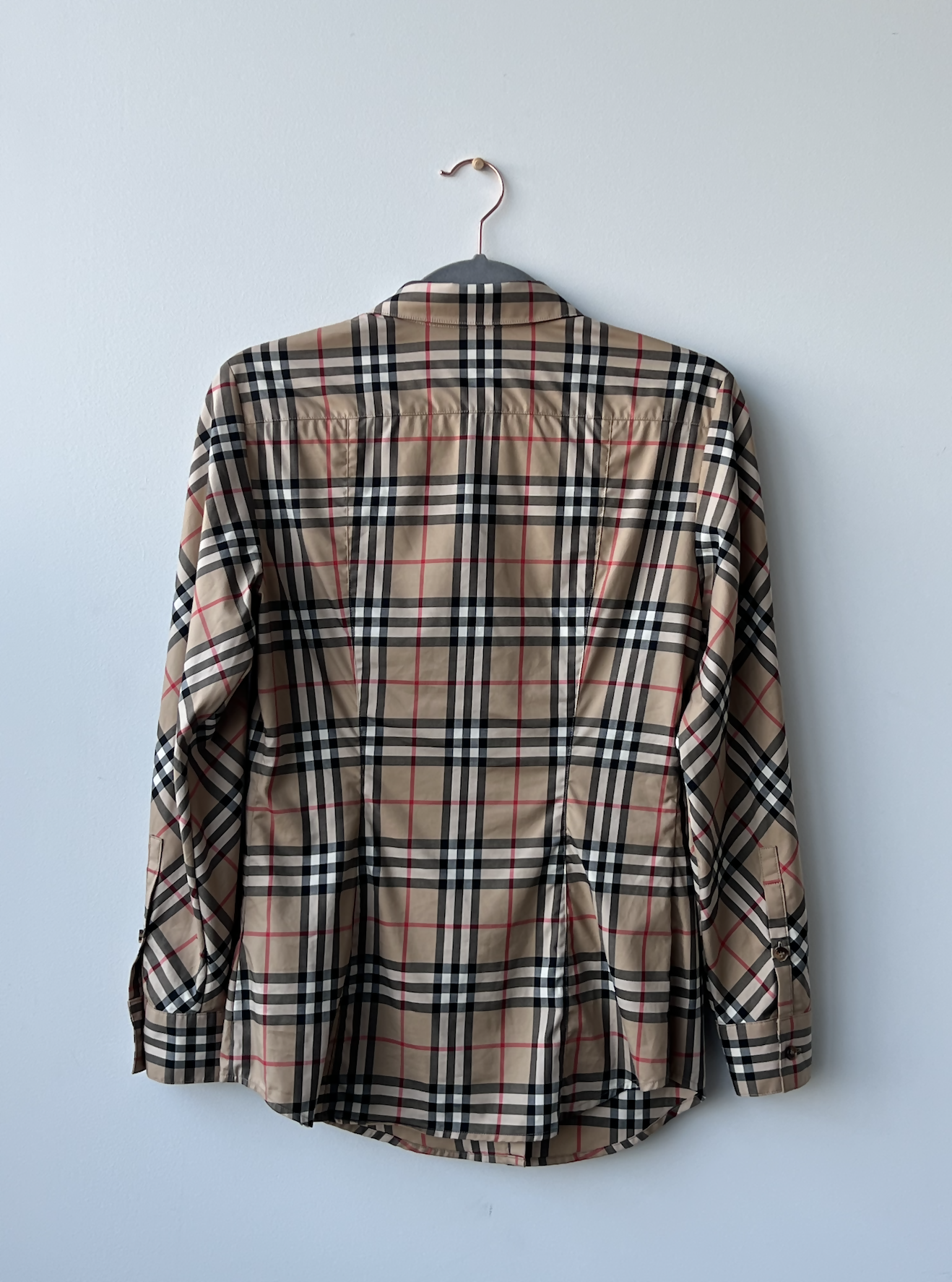 A Burrberry Button down shirt by EXCHANGE with long sleeves and classic check print hangs on a metal hanger against a light gray wall, in great condition and showing no signs of wear.