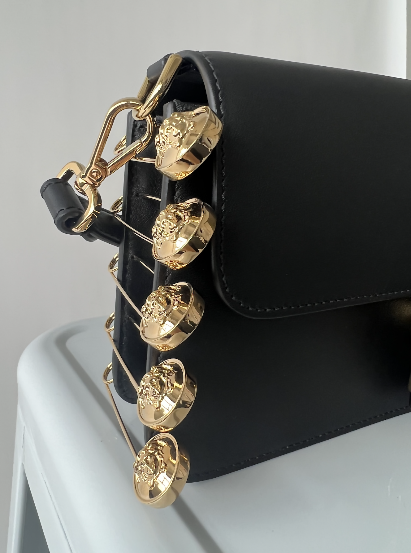 The EXCHANGE FENDI X Versace Fendace Baguette Shoulder Bag is a black leather luxury shoulder bag with a gold chain and multiple round, ornate gold charms, inspired by Italian fashion, displayed on a light gray surface.