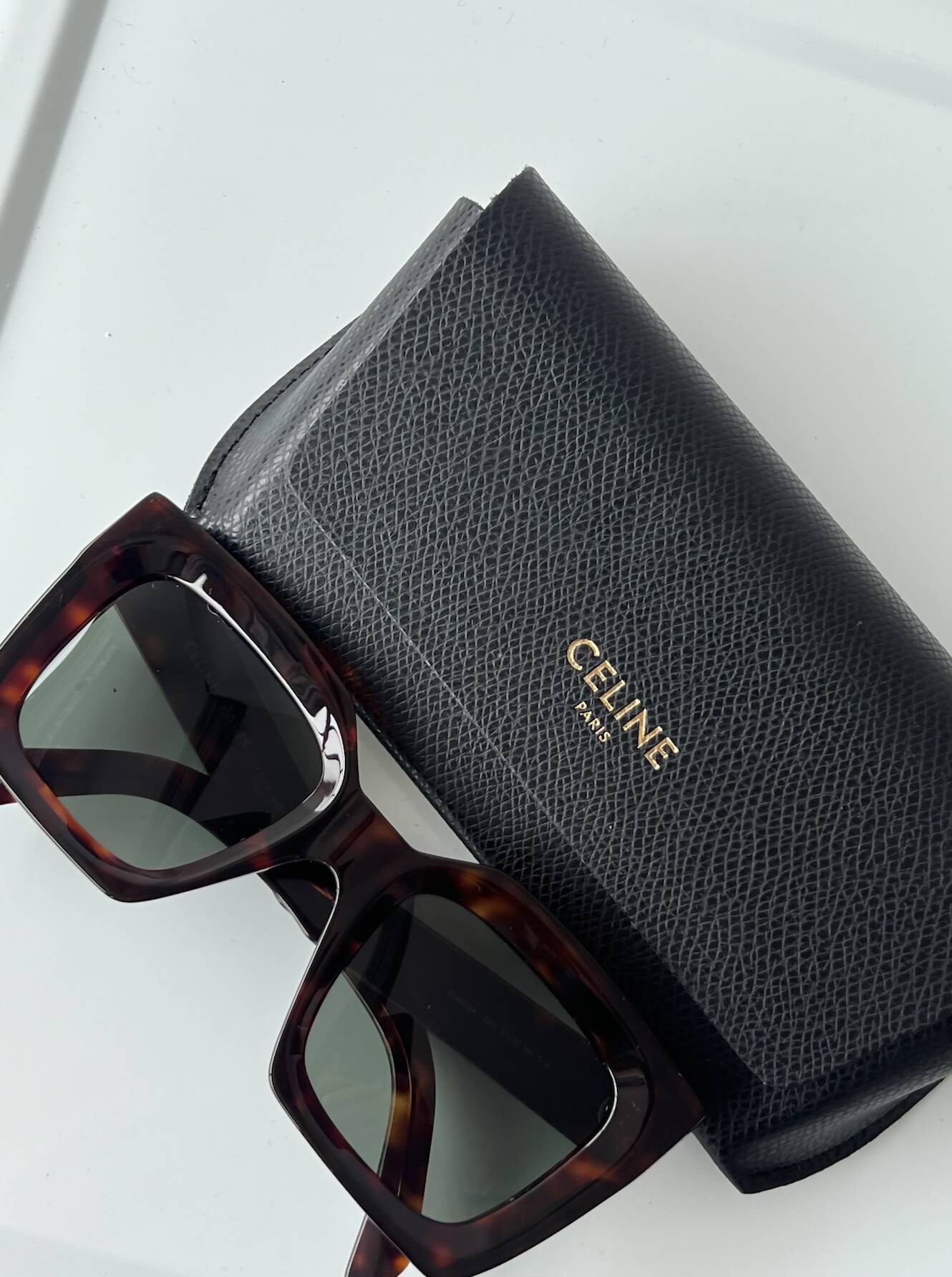 A pair of rectangular tortoiseshell Celine Glasses - CL40130I by EXCHANGE rests on a textured black case with the brand's gold logo, all set on a light-colored surface.