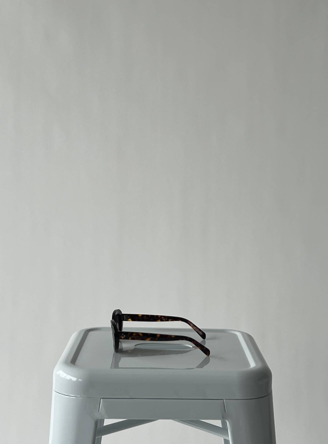 A pair of Celine Glasses by EXCHANGE with dark frames, worn once, is placed on a light gray plastic stool against a plain, light background.