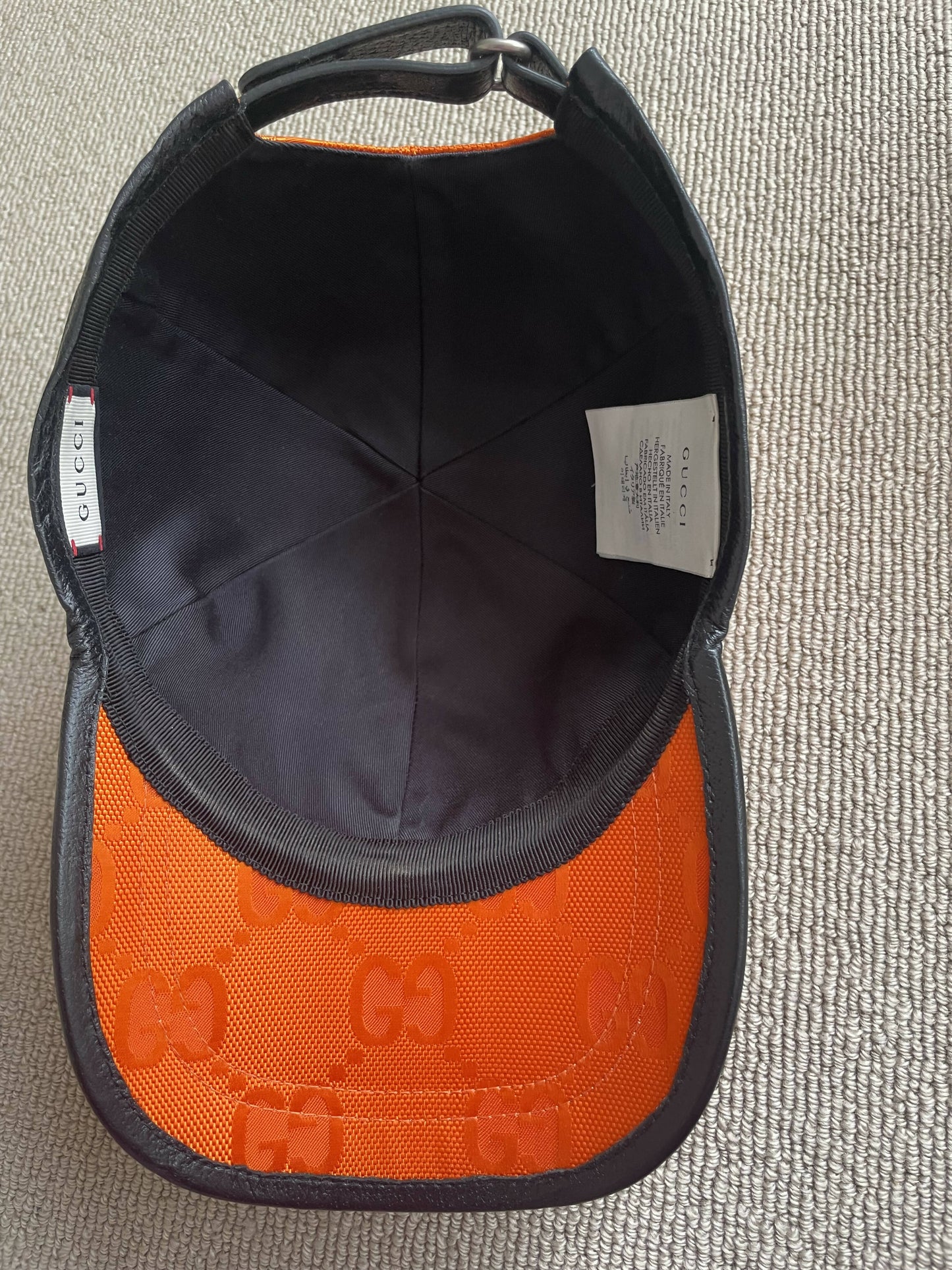 The EXCHANGE Gucci Off The Grid GG Supreme Canvas Baseball Cap in orange and black is shown from above, highlighting the inside labels and its subtle GG logo pattern on the brim, resting on a textured beige surface.