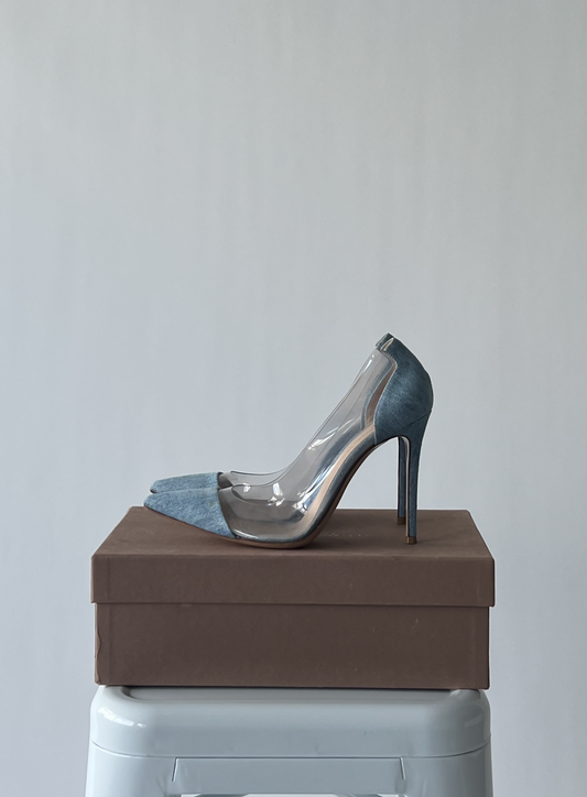 The Gianvito Rossi Light Blue Denim and PVC Plexi Pumps by EXCHANGE, in great condition, are displayed atop a closed brown shoebox on a white stool against a plain light gray background.