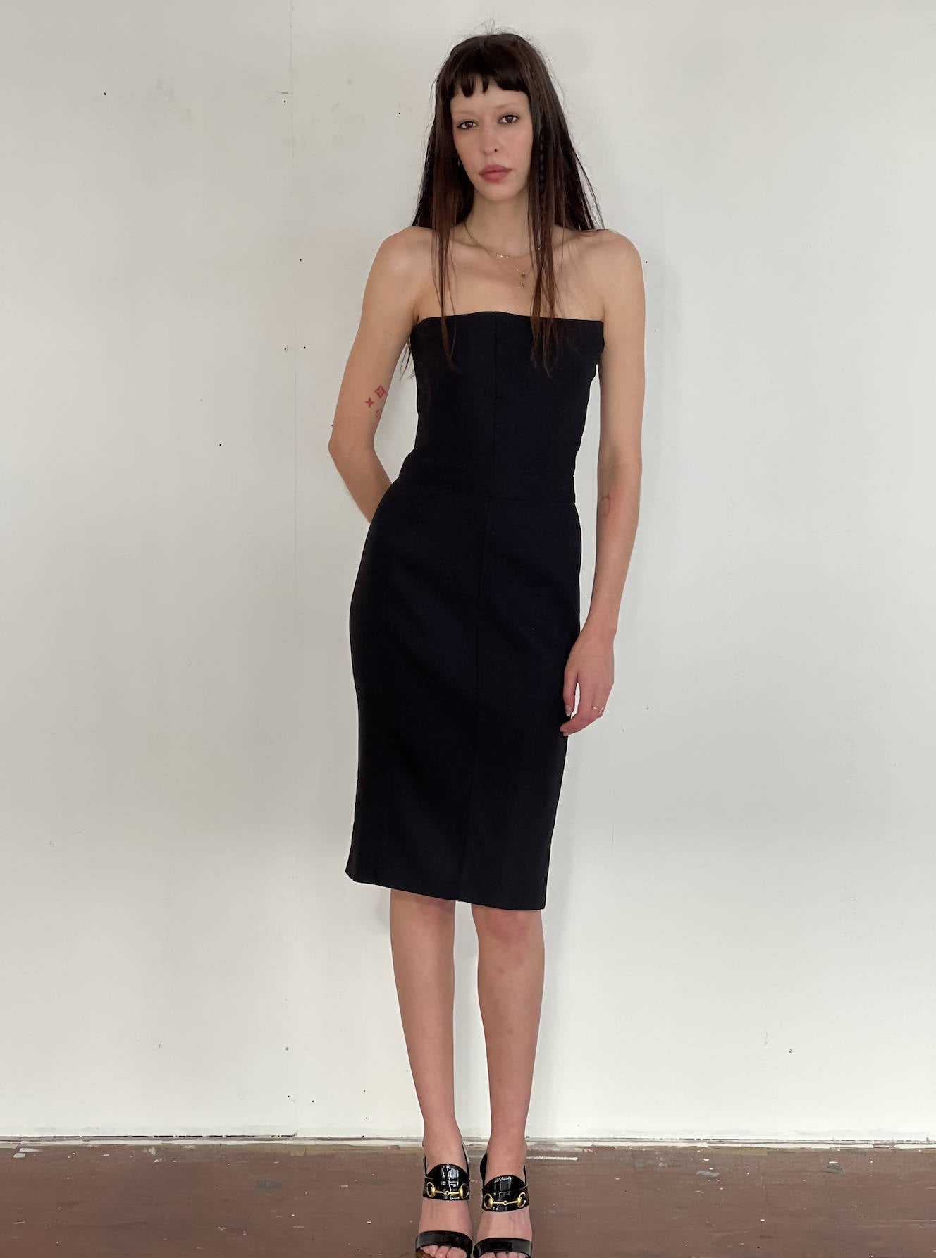 A woman with long dark hair wears the EXCHANGE Stella McCartney Black Dress and black open-toe heels, standing against a plain white wall and wooden floor.