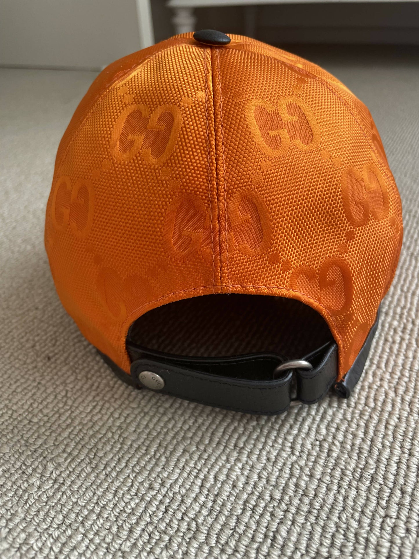 The Gucci Off The Grid GG Supreme Canvas Baseball Cap by EXCHANGE, in orange with a repeating GG pattern, features a black adjustable leather strap and rests on a light-colored carpeted floor.