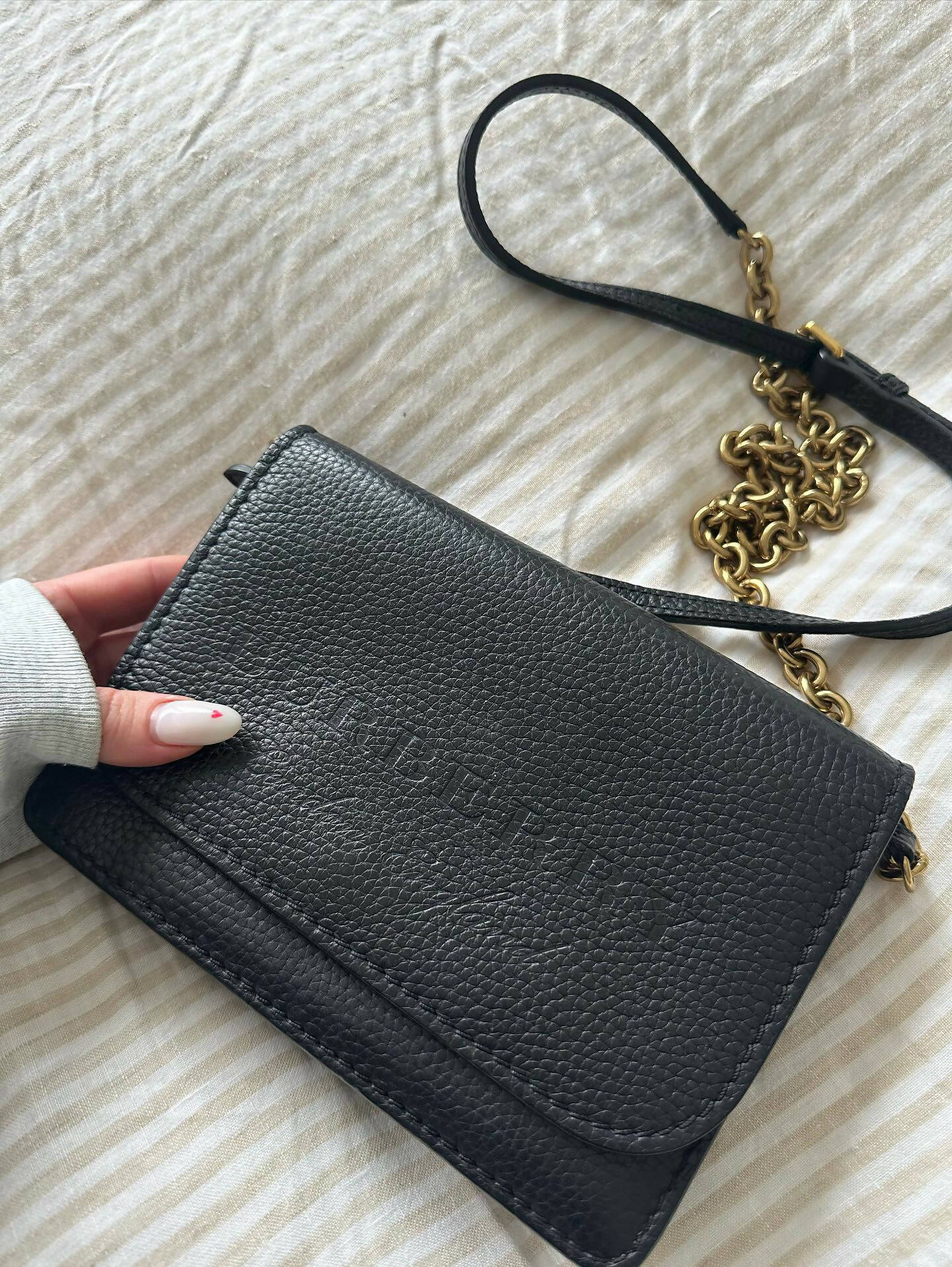 A hand holds a black grained leather EXCHANGE Burberry Logo Embossed Wallet On Chain with gold hardware and chain strap, resting on a light-colored striped fabric surface.
