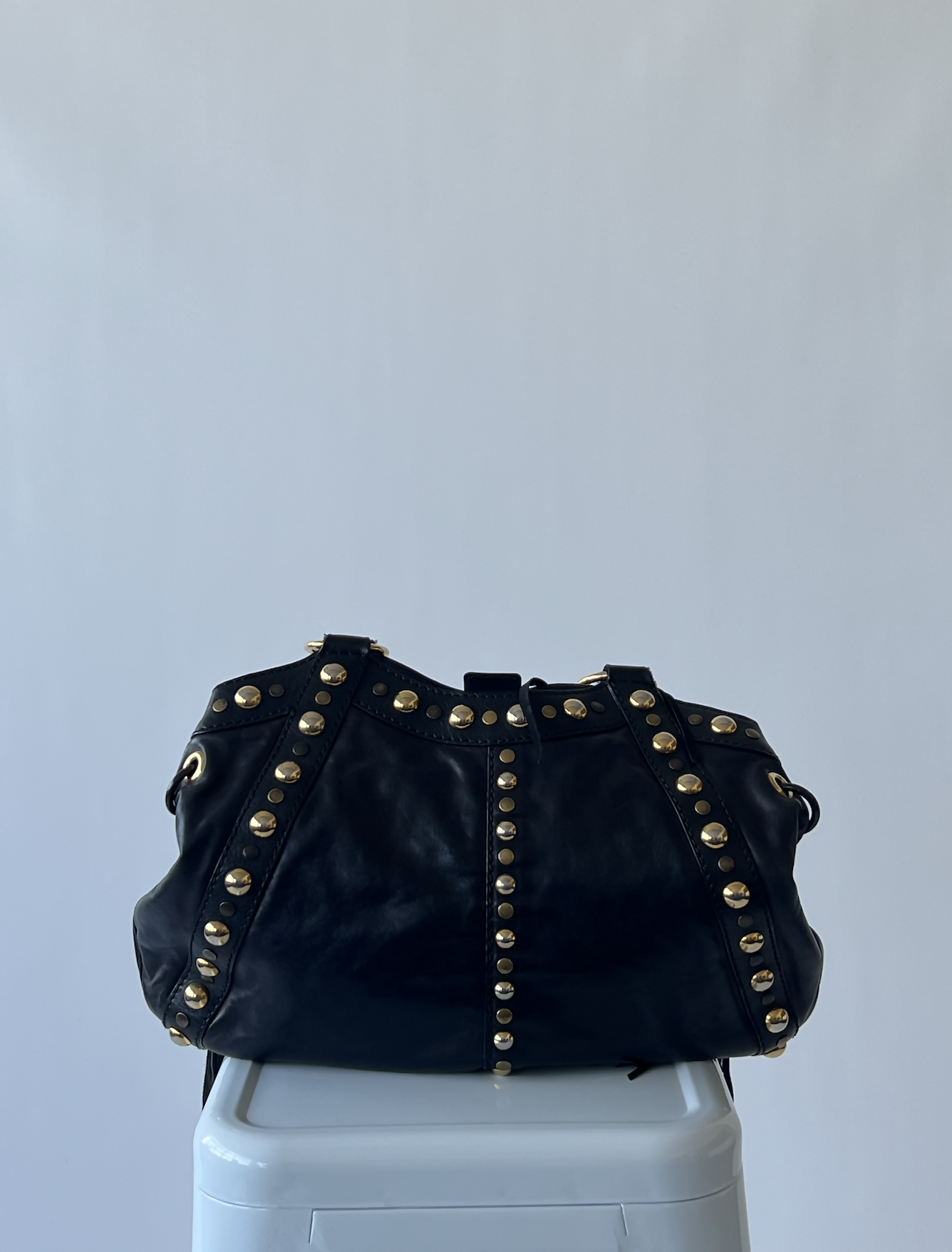 The Vintage Gucci Bag by EXCHANGE, crafted from black leather and embellished with rows of gold studs, is displayed on a white pedestal against a plain light gray background.
