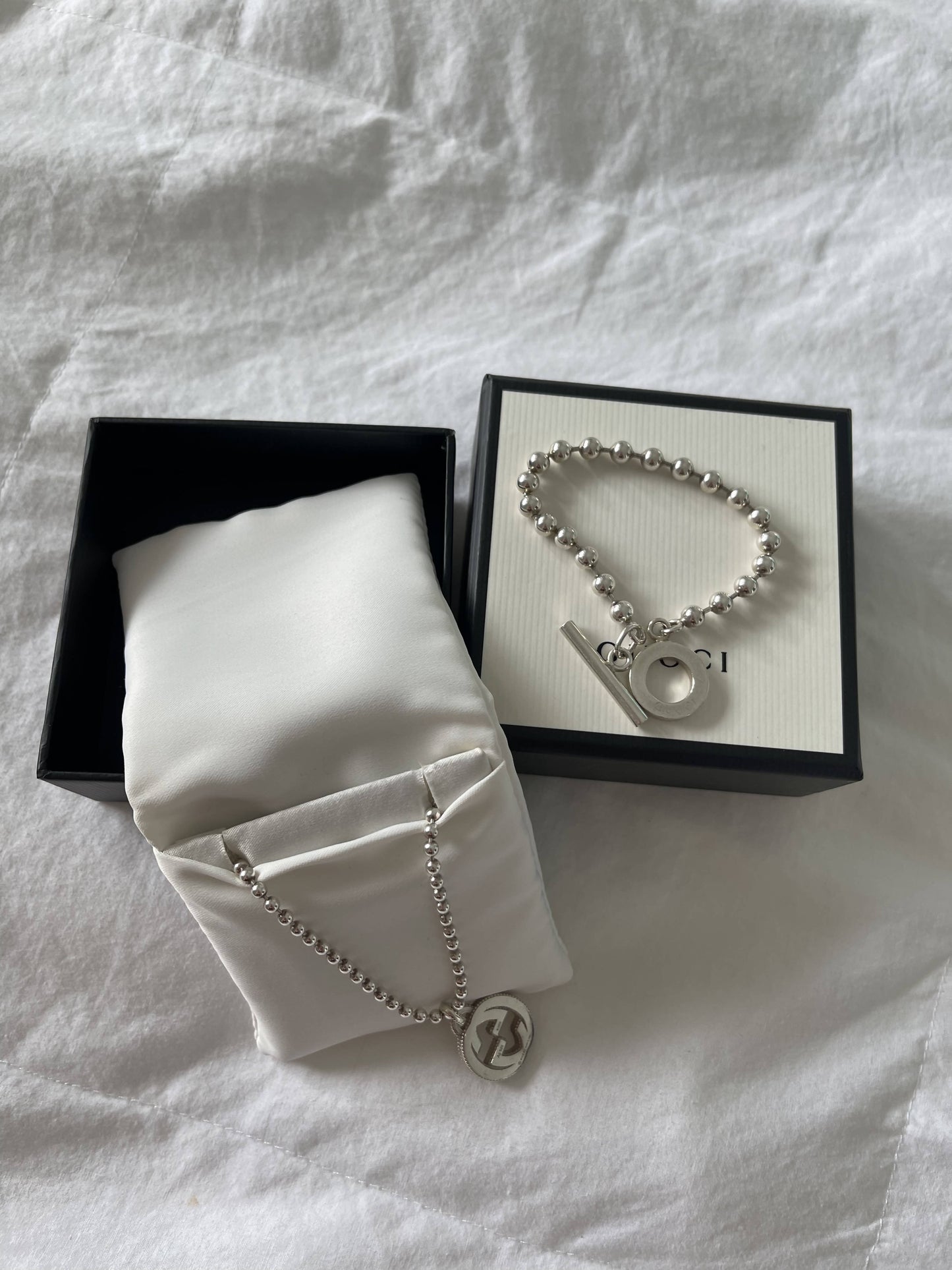 The EXCHANGE Gucci Bracelet, featuring round silver beads and a toggle clasp, sits on a cream box next to a matching necklace displayed on white satin inside a black jewelry box, all atop white fabric.