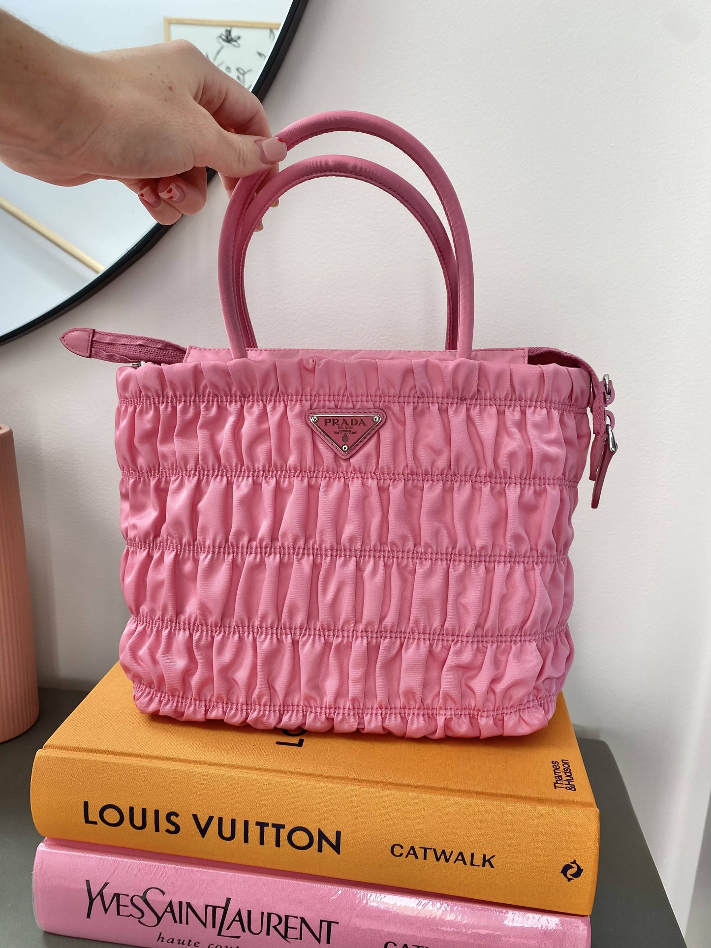 A hand holds an EXCHANGE Prada Pink Nylon Tote atop two fashion books—Louis Vuitton Catwalk and Yves Saint Laurent Catwalk—on a gray surface by a mirror.