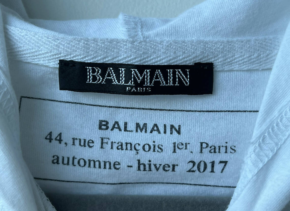 Close-up of a white clothing label inside an EXCHANGE Balmain Long Sleeve Hoodie 2017, featuring a black brand tag, address (44 rue François 1er, Paris), and "autumn - winter 2017." This hoodie is made from 100% cotton.