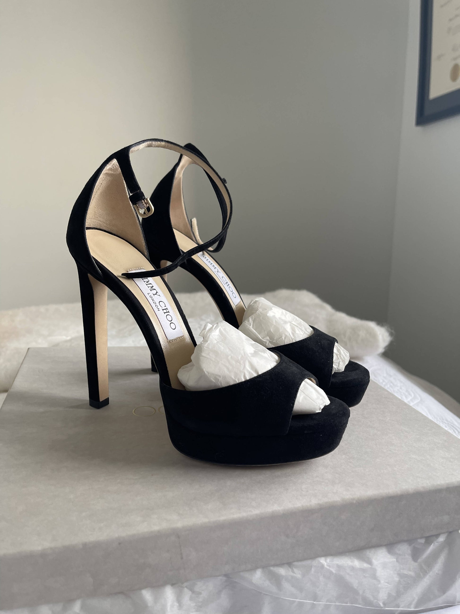 A pair of black Jimmy Choo Pattie Heels by EXCHANGE in size 37.5 with ankle straps, shown on a light-colored box with tissue paper inside; these sandals are in excellent condition.