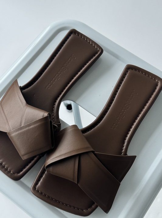 The ACNE STUDIOS MUSUBI SANDAL by EXCHANGE features brown, square-toed slides with wide, knotted straps inspired by the iconic Musubi knot and crafted from soft calf leather, displayed on a white square chair.