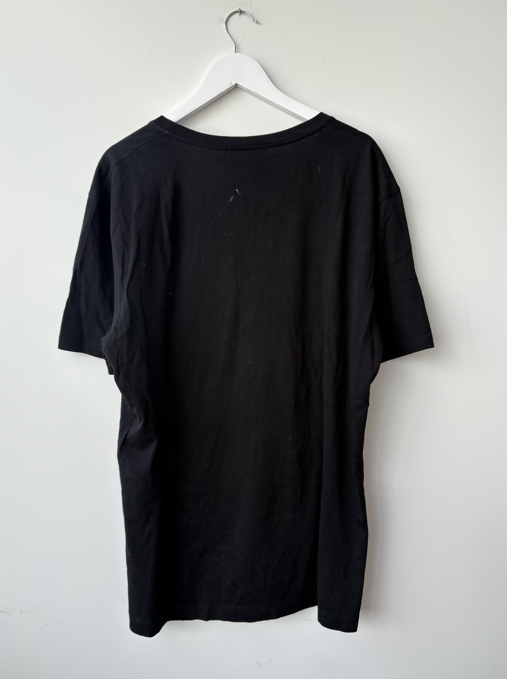 The Gucci Black L'Aveugle Par Amour T-Shirt from EXCHANGE STORE hangs on a white plastic hanger against a white wall, displayed from the back to highlight its minimalist luxury style.