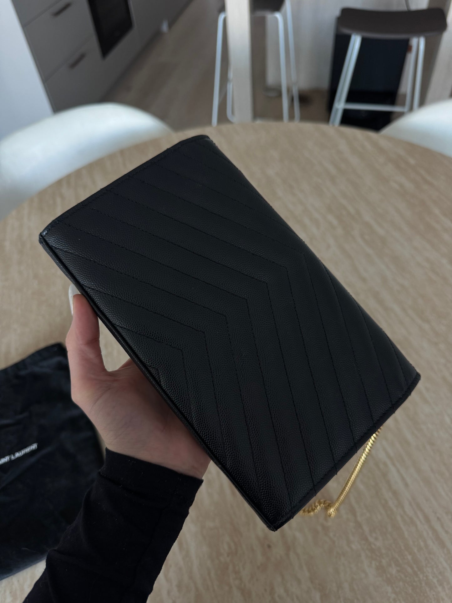 A hand holds the EXCHANGE STORE Saint Laurent Classic Cassandre chain wallet in black grain de poudre leather with a gold chain strap, over a light wooden table. Chairs and part of a kitchen appear in the background.