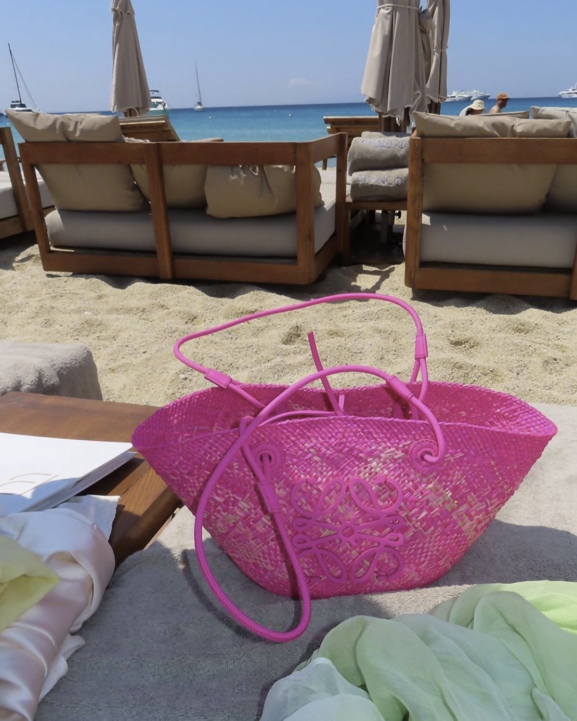 The Loewe Raffia Pink Wicker Basket Tote 2023 by EXCHANGE STORE sits in great condition on beach lounge chairs, with umbrellas, the sea, and anchored boats visible under a clear blue sky.