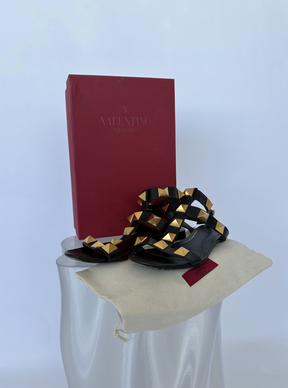The EXCHANGE STORE Valentino Garavani Roman Stud Sandals Black - FULL SET features black leather sandals with gold pyramid studs, presented on a cream dust bag and displayed in front of a red Valentino box against a light background.