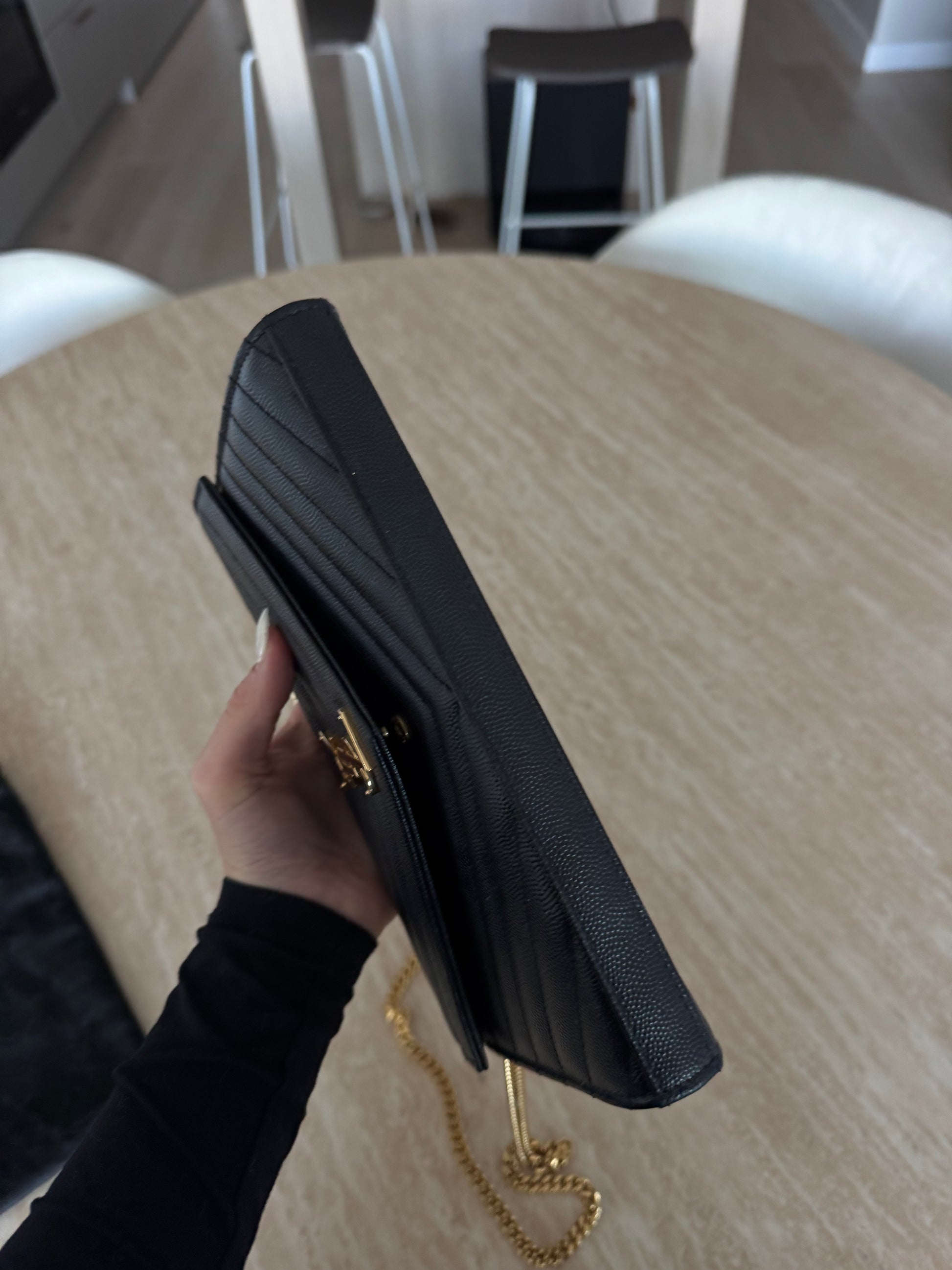 A person holds the EXCHANGE STORE Saint Laurent Classic Cassandre chain wallet in black grain de poudre leather over a round wooden table, with white chairs in the background.